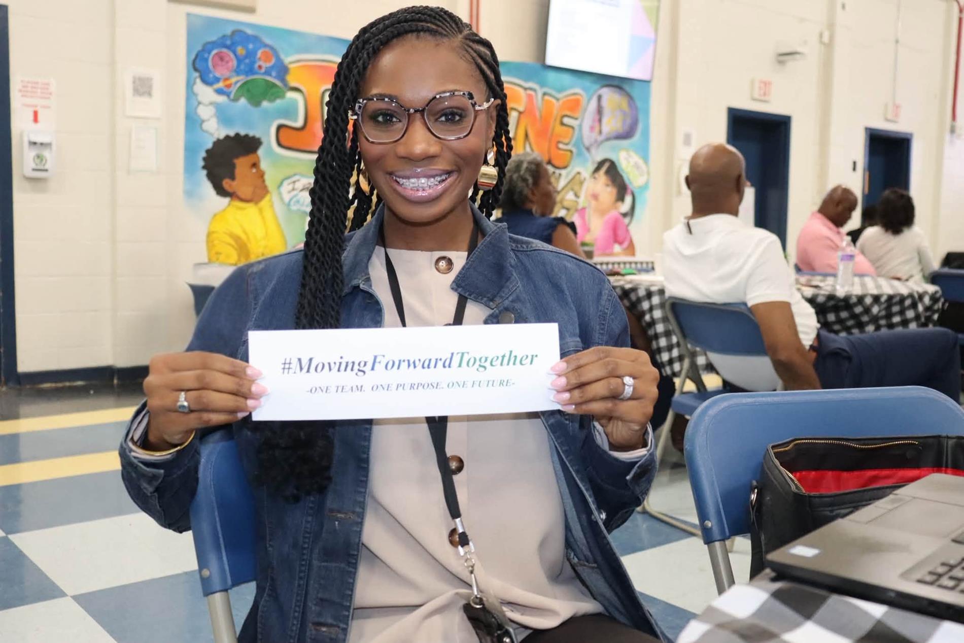 A smiling Williamsburg County School District staff member wearing glasses and a denim jacket sits at a table during a Summer Leadership event, holding a card that reads “#MovingForwardTogether – One Team. One Purpose. One Future.” Other attendees and a colorful educational mural are visible in the background.