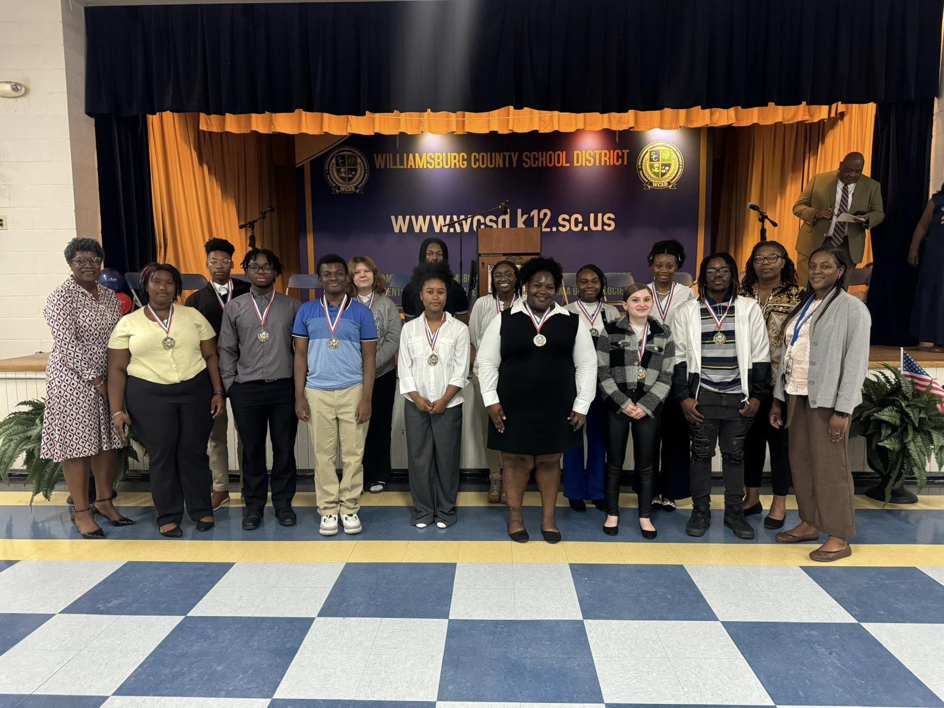 A group of high school students stand in front of a stage with a “Williamsburg County School District” banner and website displayed behind them. The students wear school uniforms or casual outfits and have medals around their necks, posing in a line. Several adults stand alongside them, one holding a trophy. The stage features gold curtains, chairs, microphones, and a podium, indicating an awards or recognition ceremony.