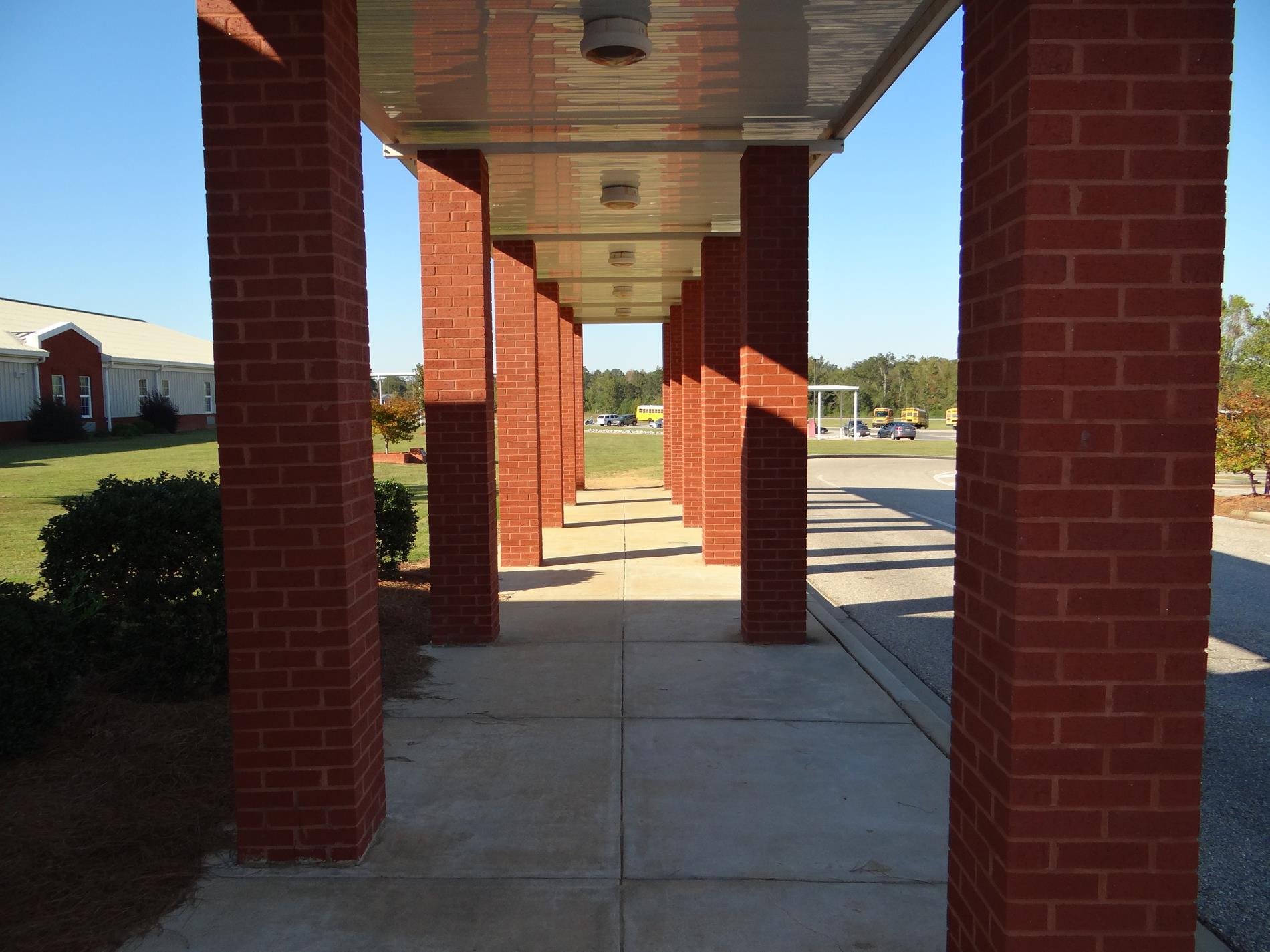 View from the front doors of Clanton Middle School to the bus road - a sidewalk covered by a white metal awning that is supported by small brick columns and framed by a row of holly bushes