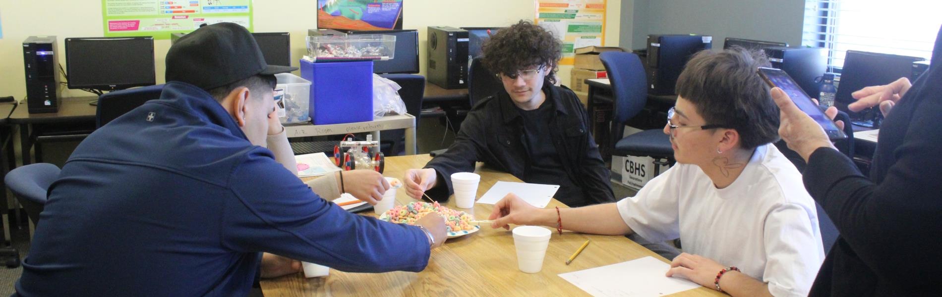 Four students sit at a table picking up cereal with objects such as a straw, toothpick, clothespin and spoon, while a teacher times them.