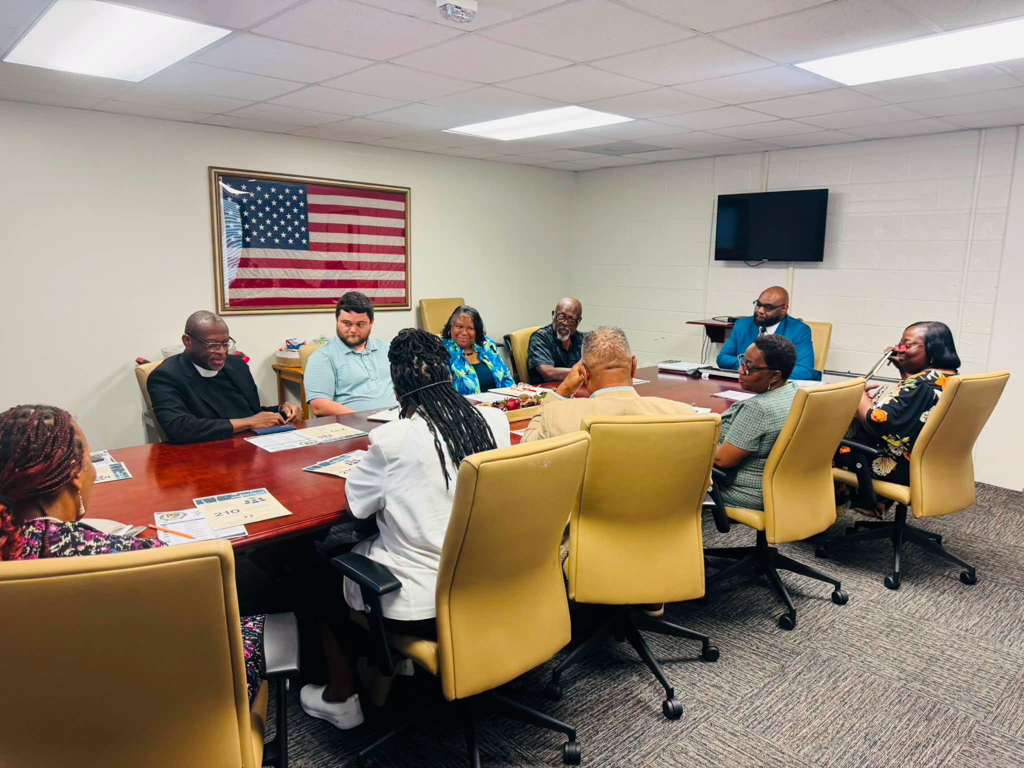 group of people seated around a large conference table. visible in the background is an American flag and a television