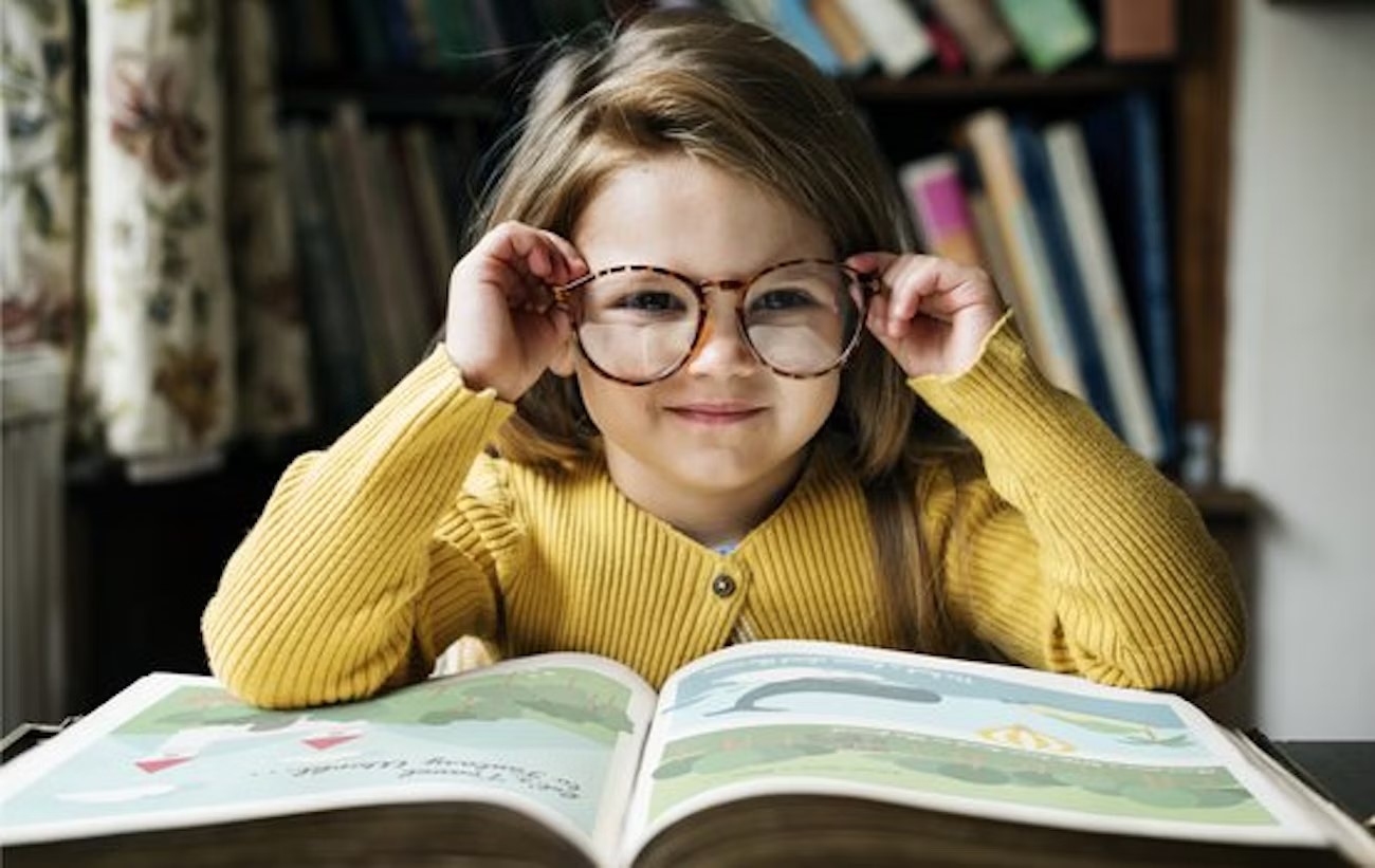 Little girl with brown bob hair cut, in a yellow sweater, touching her dark black glasses
