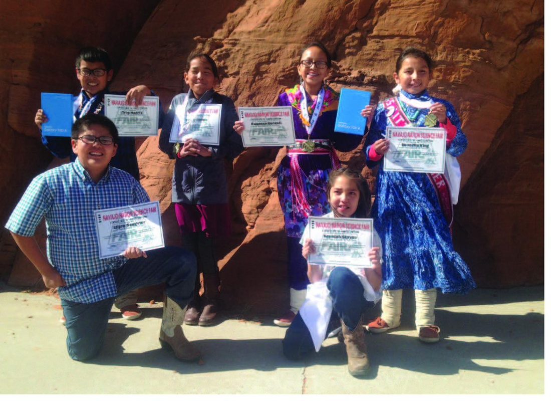 Students in traditional Diné attire holding science-fair award certificates at red sandstone cliffs.