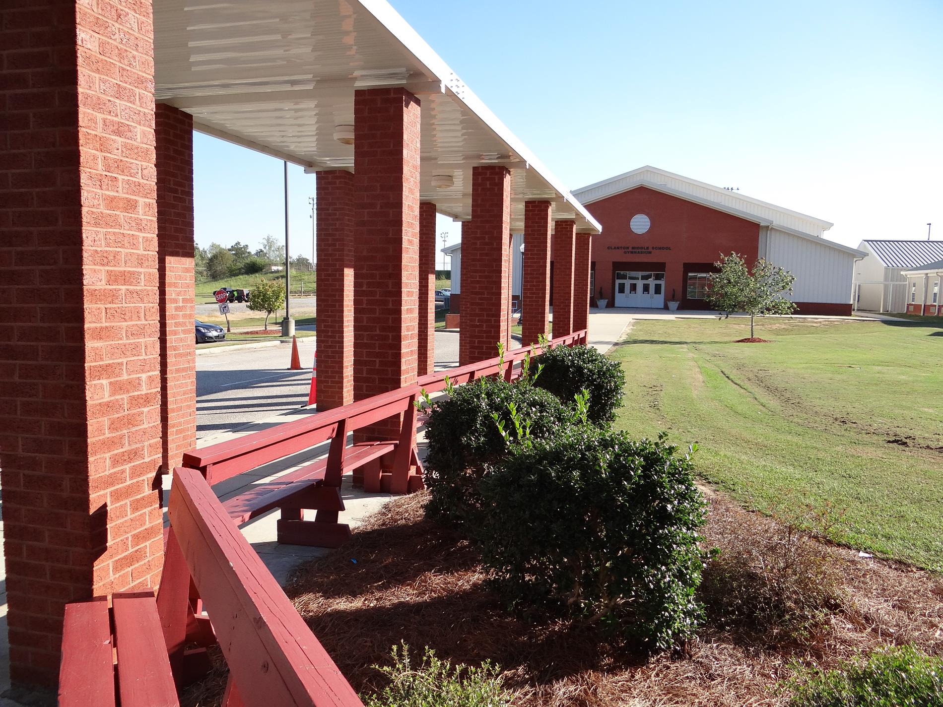 View from the front doors of Clanton Middle School to the gym - a sidewalk covered by a white metal awning that is supported by small brick columns and framed by a row of red benches with a row of holly bushes behind