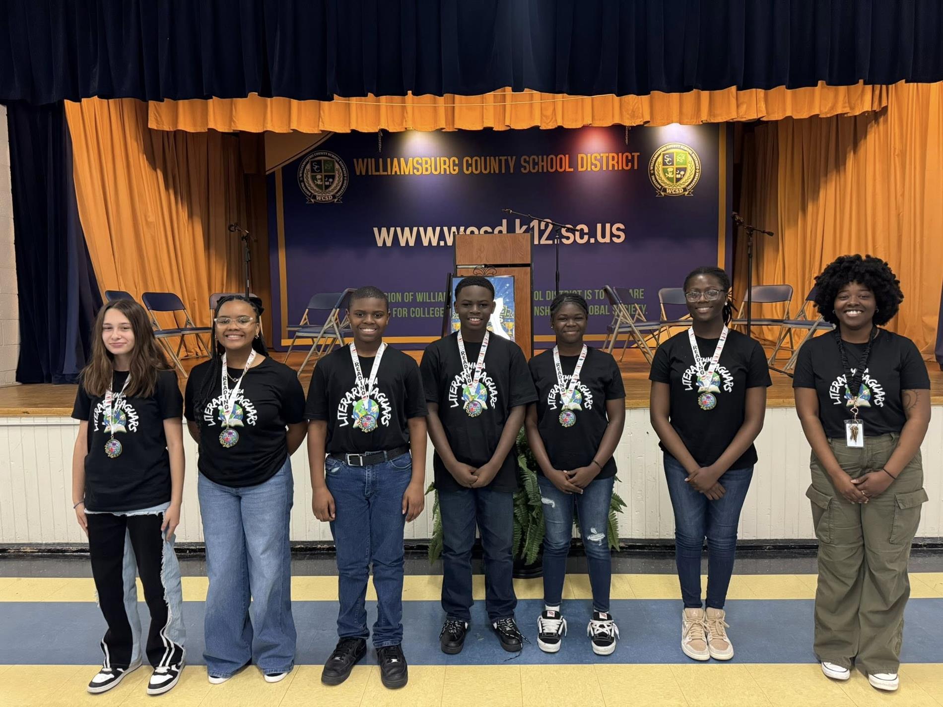 students and one adult standing in front of a blue and orange curtained backdrop with a banner that reads "WILLIAMSBURG COUNTY SCHOOL DISTRICT" and the district's website, "www.wcsd.k12.sc.us." 