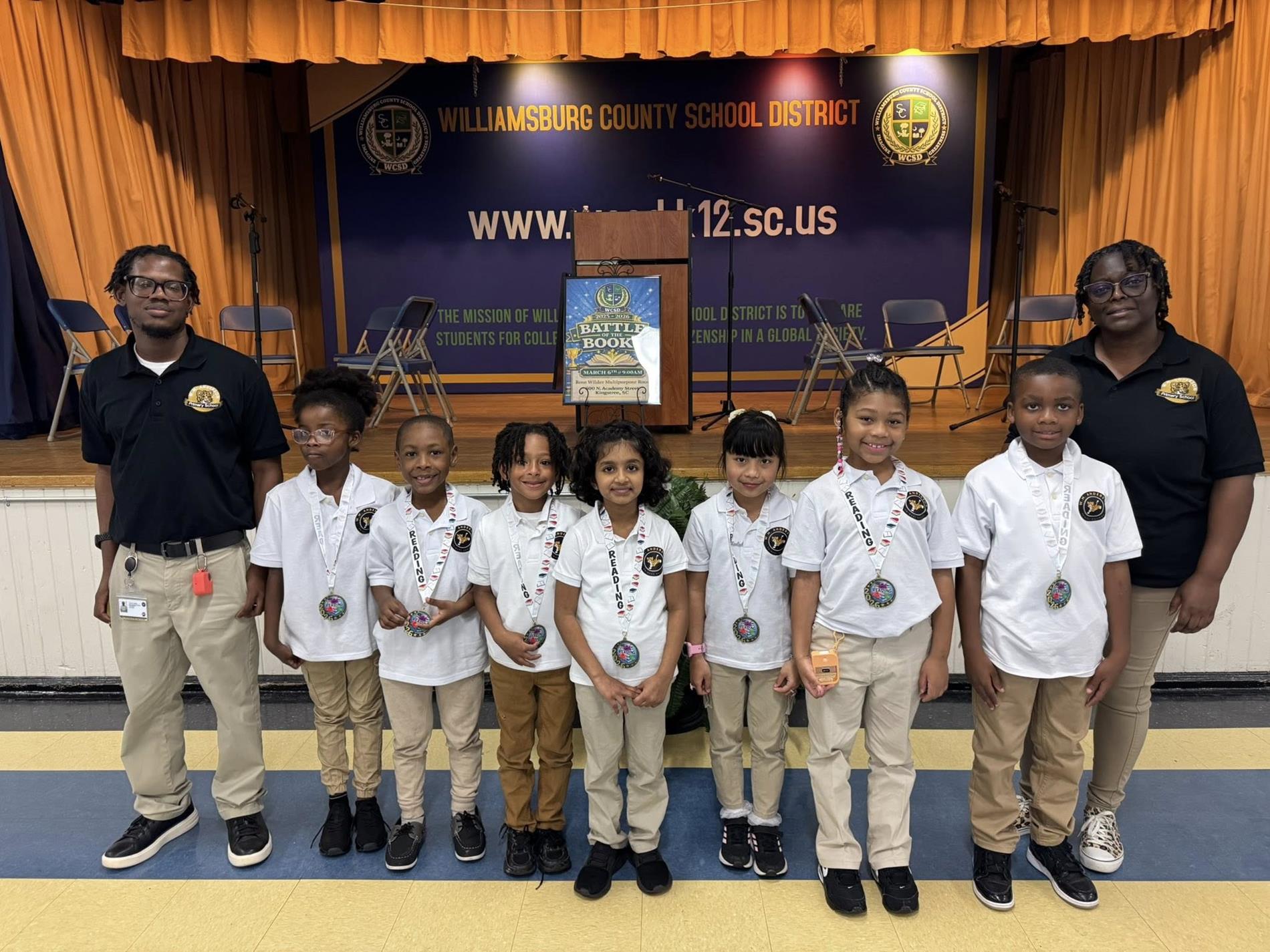 Six young students wearing white polo shirts, khaki pants, and medals with "READING" ribbons, pose for a photo on a stage with two adult chaperones in matching black polo shirts. The stage features orange curtains and a banner with "WILLIAMSBURG COUNTY SCHOOL DISTRICT" and their website, "www.wcsd.k12.sc.us." A poster for "BATTLE OF THE BOOKS" is in the background.
