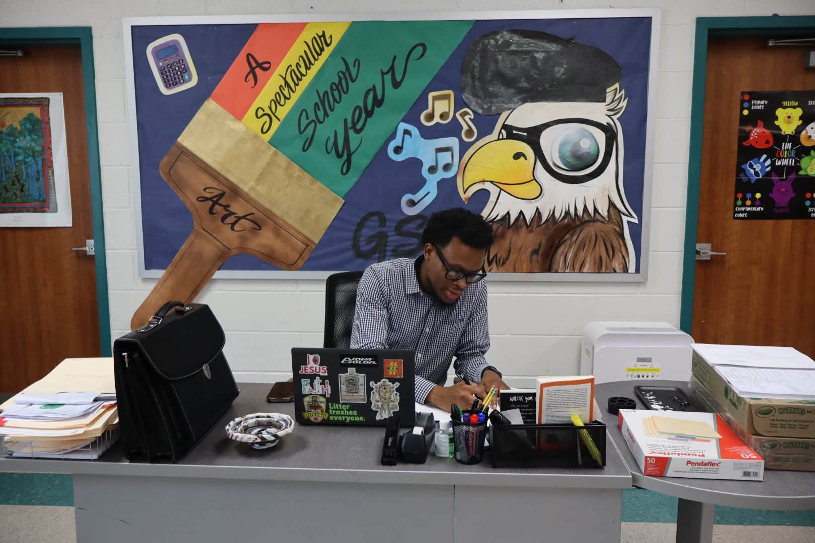 A man wearing glasses and a checkered shirt sits at a school office desk, writing on paperwork beside a laptop covered with stickers. The desk is surrounded by folders, office supplies, a briefcase, printer, and storage boxes. Behind him, a colorful mural reads “A Spectacular School Year” and features art and music imagery alongside a large cartoon eagle.