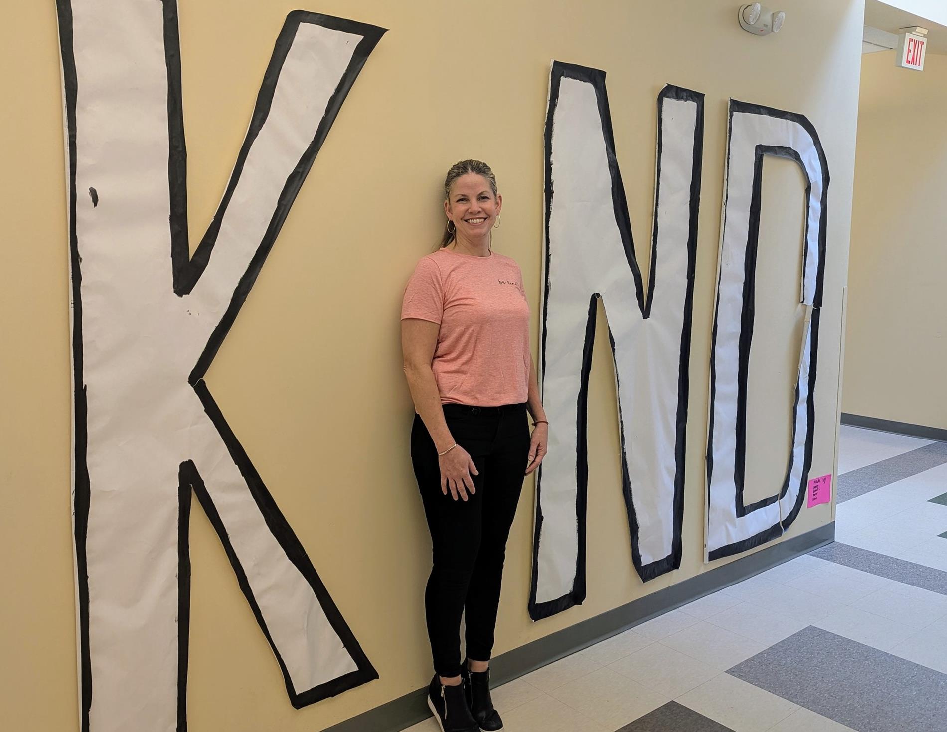 Director of Student Services wearing a pink shirt. The director stands agains a the wall, forming the "i" in a large sign reading "KIND".