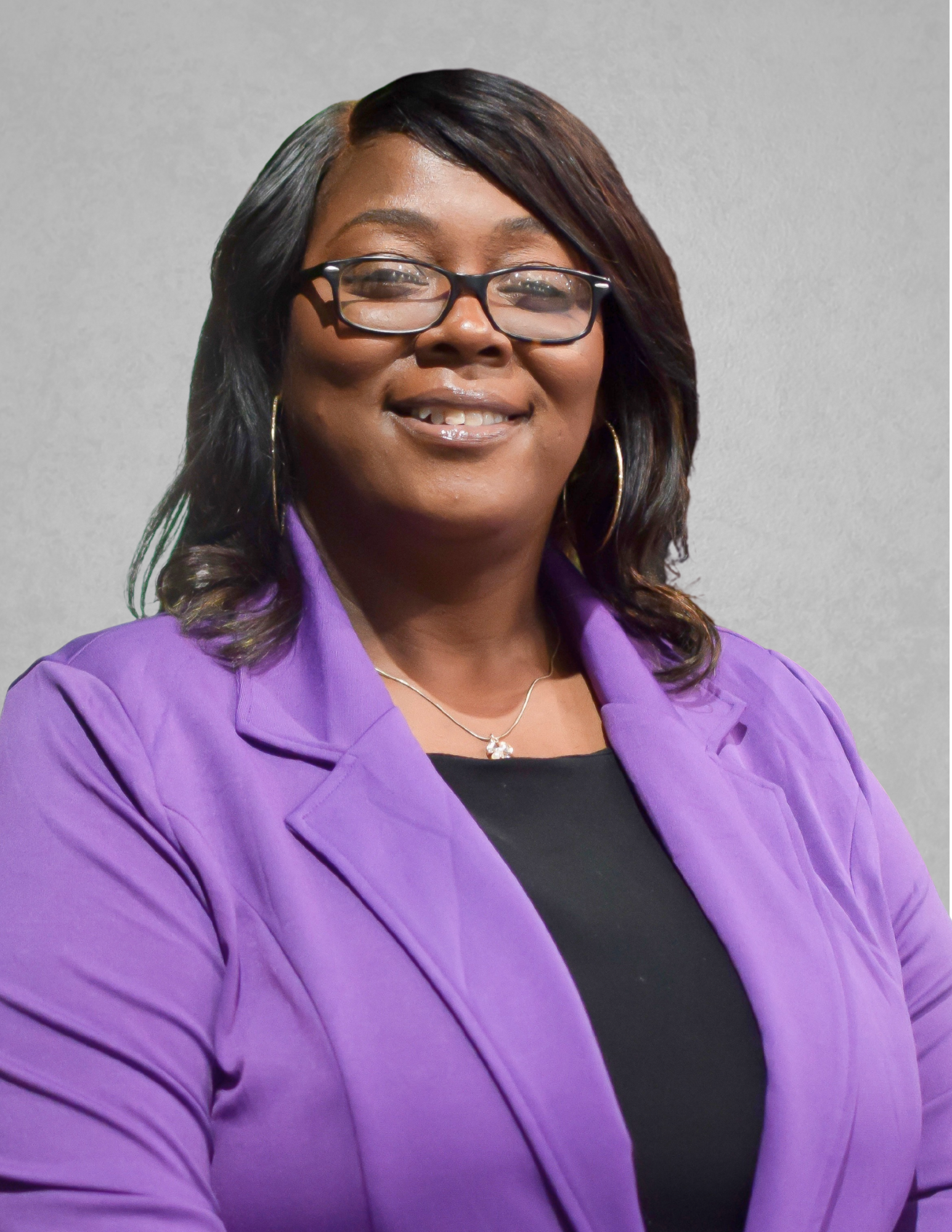 rofessional portrait of Claudine Rogers smiling and facing the camera. She is wearing glasses, a purple blazer over a black top, hoop earrings, and a silver necklace. Rogers has shoulder-length dark hair and is posed against a light gray background.