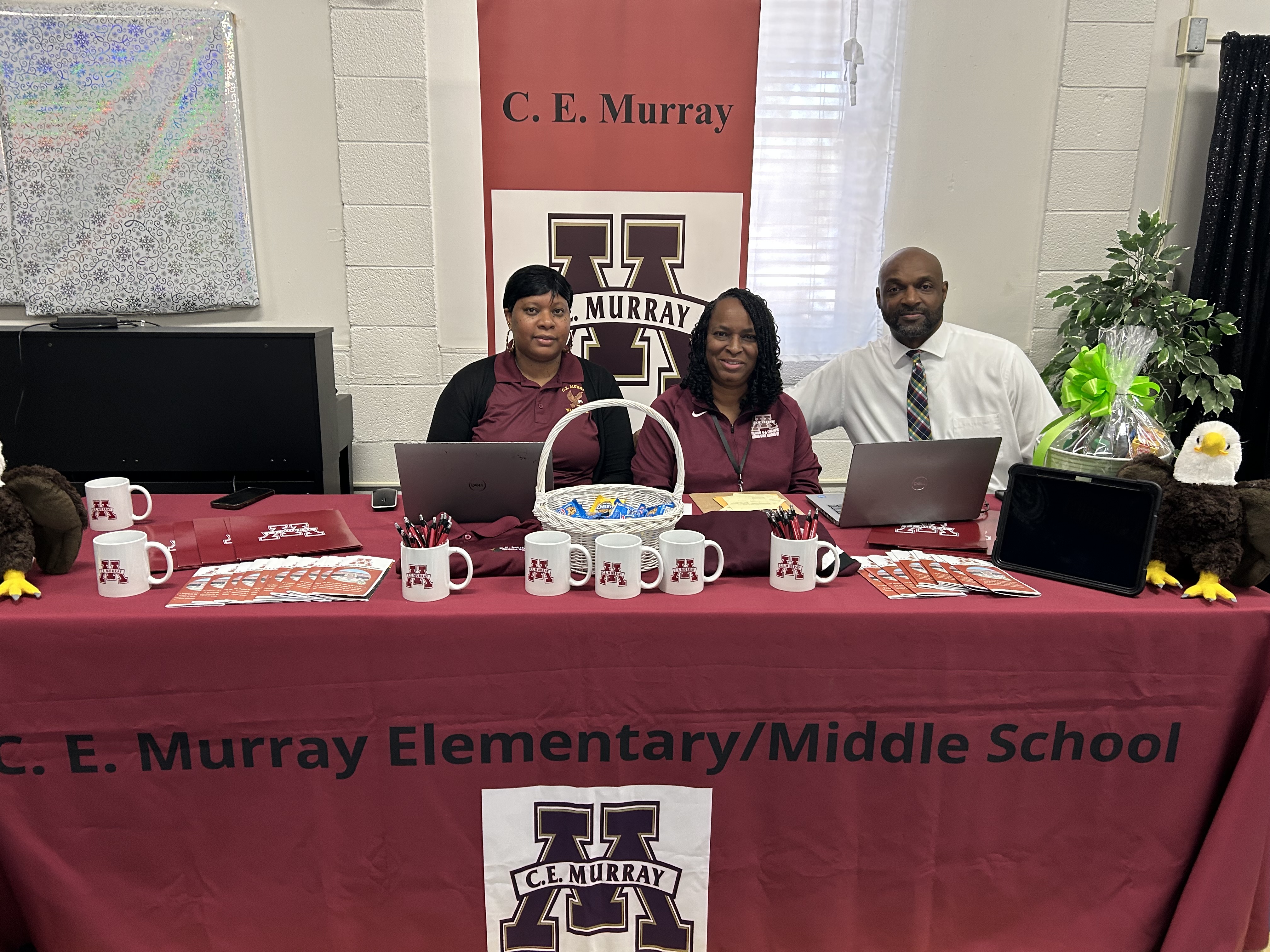 Alt text: Three staff members sit behind a maroon table at a C. E. Murray Elementary/Middle School display. A banner behind them reads “C. E. Murray” with a large “M” logo. The table is covered with school-branded items including mugs, pens, brochures, laptops, a basket of snacks, and two eagle mascots on either end.