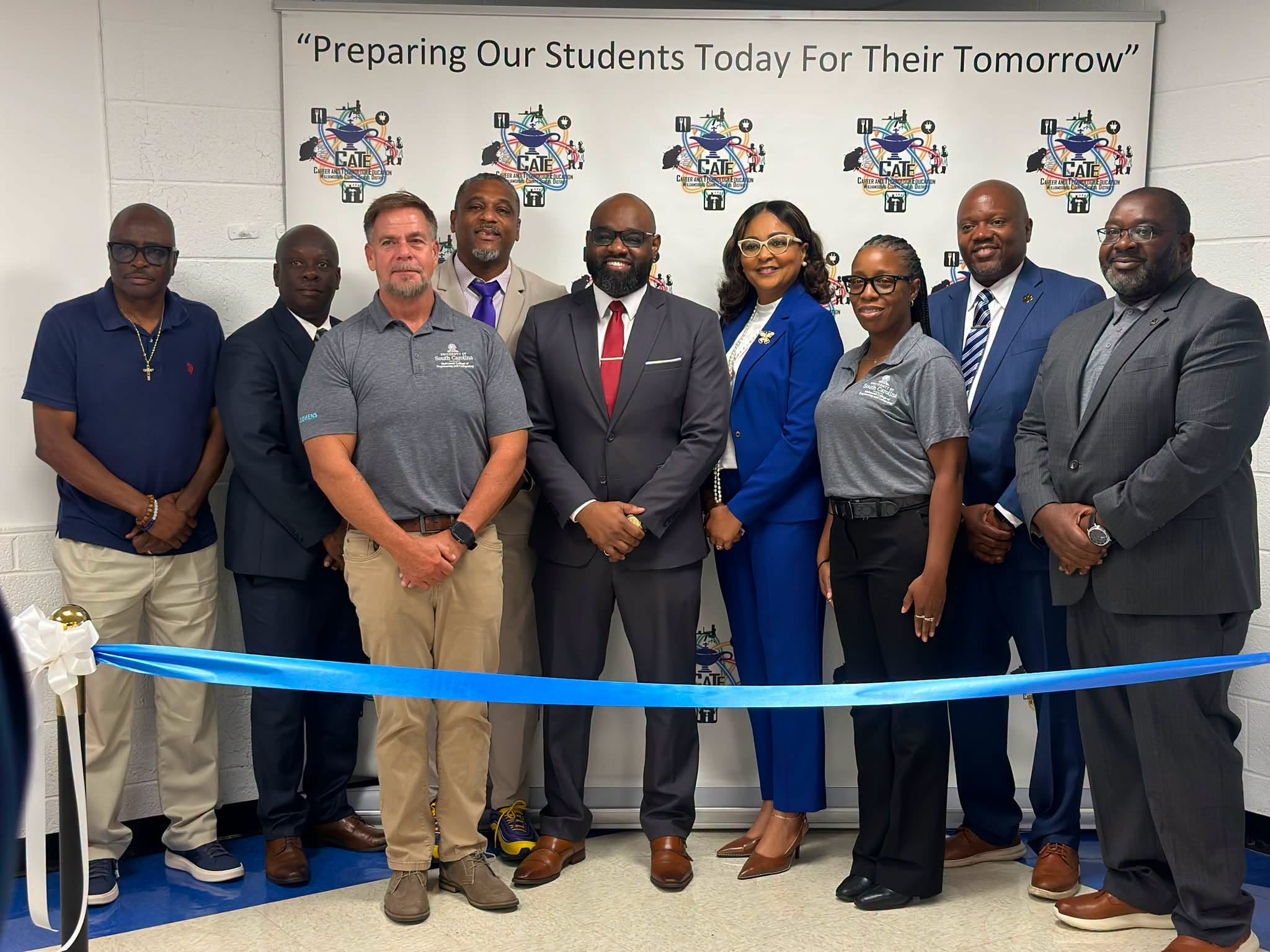A group of nine school and community leaders pose together behind a blue ceremonial ribbon at a Career and Technology Education event. They stand in front of a CATE backdrop displaying the message, “Preparing Our Students Today For Their Tomorrow.”
