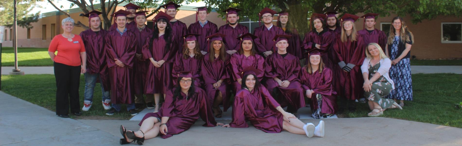 Twenty-One Graduates in Maroon Caps and Gowns pose by a bench in three rows one row standing, one row sitting on a bench, two girls on the ground reclining. They are flanked by three teachers. They are posed under multiple trees in front of a light colored brick high school building.