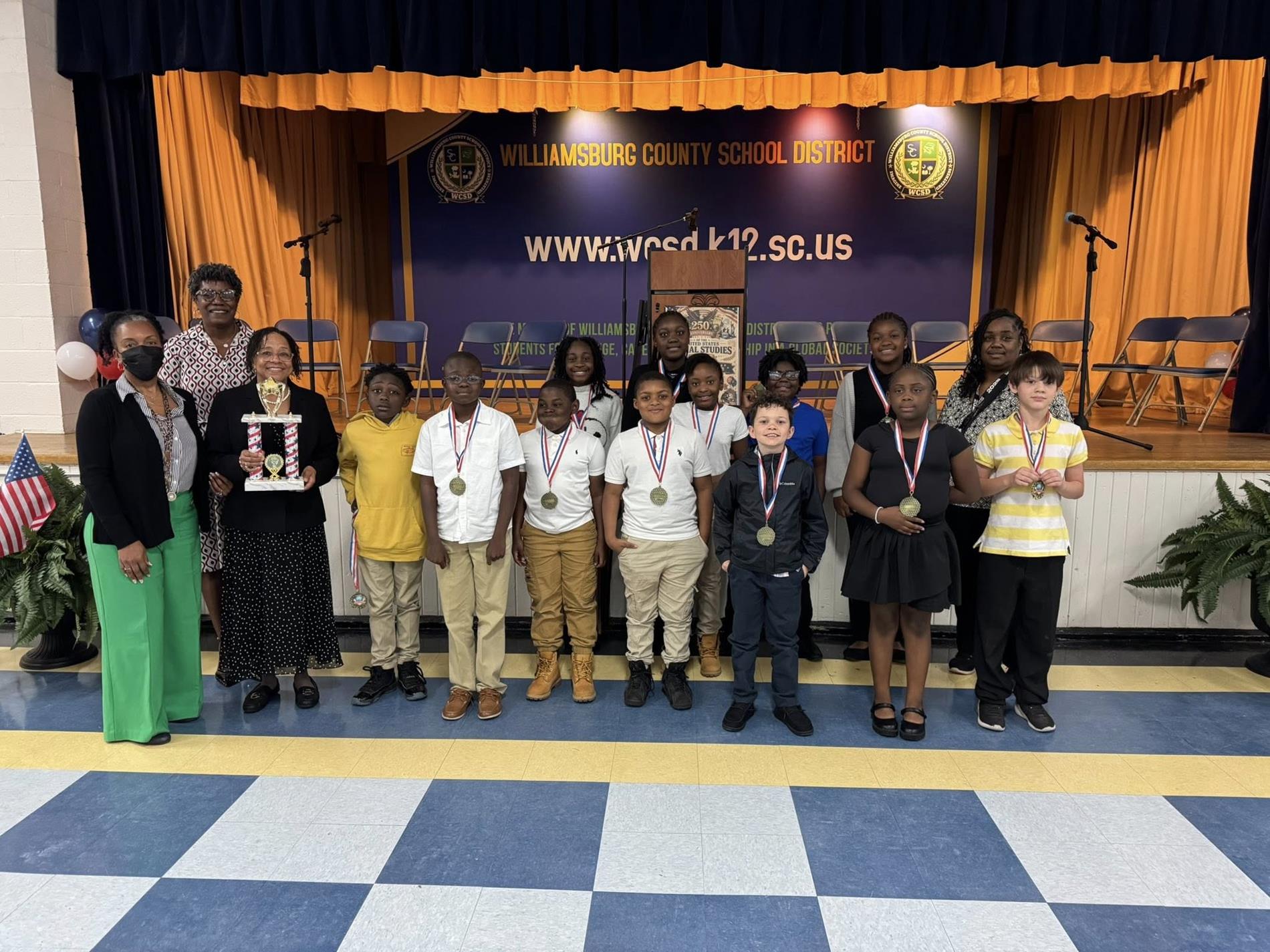 A group of elementary school students stand in front of a stage with a “Williamsburg County School District” banner and website displayed behind them. The students wear school uniforms or casual outfits and have medals around their necks, posing in a line. Several adults stand alongside them, one holding a trophy. The stage features gold curtains, chairs, microphones, and a podium, indicating an awards or recognition ceremony.