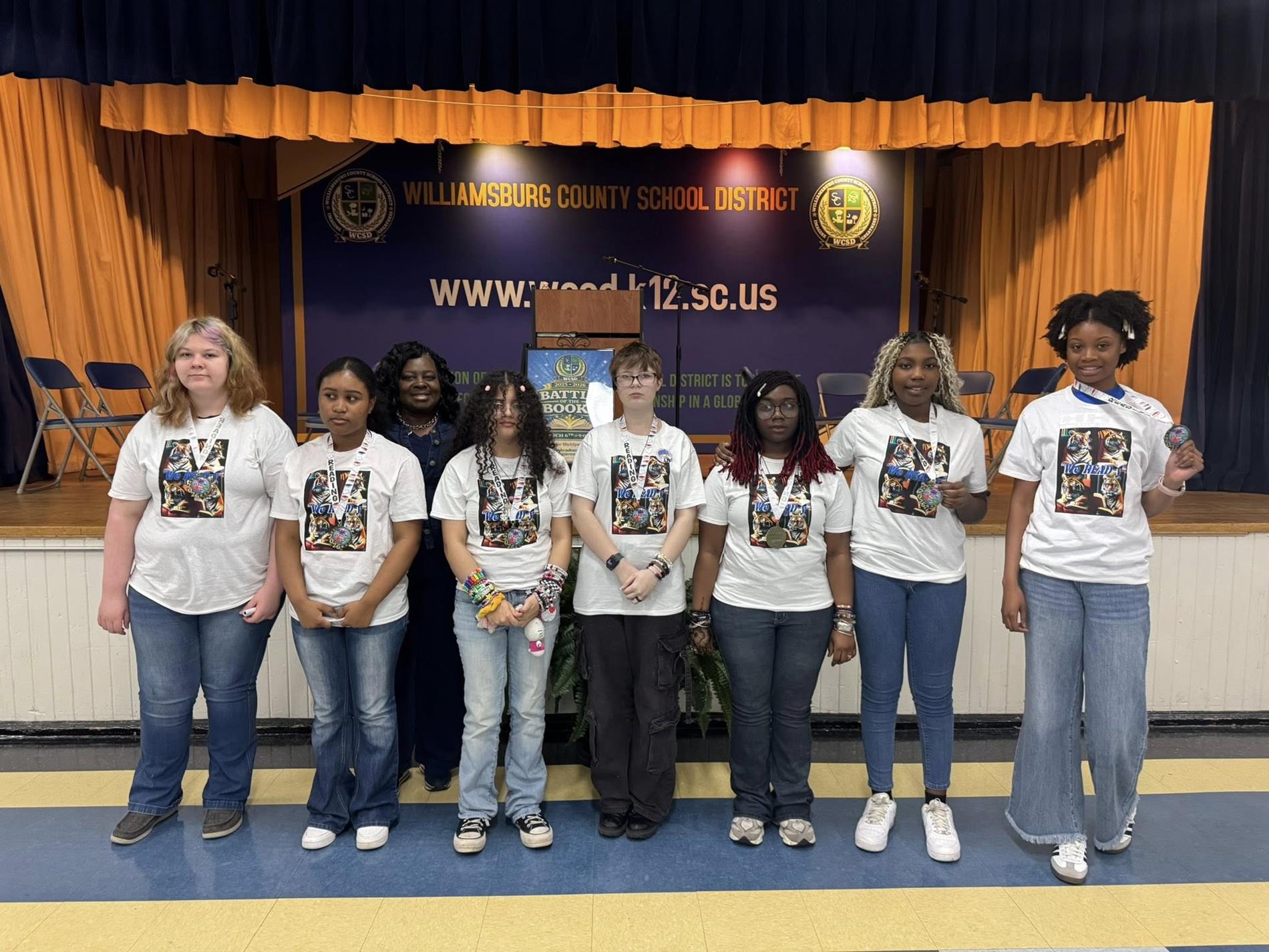 A group photograph featuring five students and a woman standing in front of stage, posing for the camera. The students, all female, are positioned in the front row and are wearing white short-sleeved t-shirts with a graphic print. stage with a blue and yellow striped floor, in front of a blue and orange curtained backdrop with a banner that reads "WILLIAMSBURG COUNTY SCHOOL DISTRICT" and the district's website, "www.wcsd.k12.sc.us." 