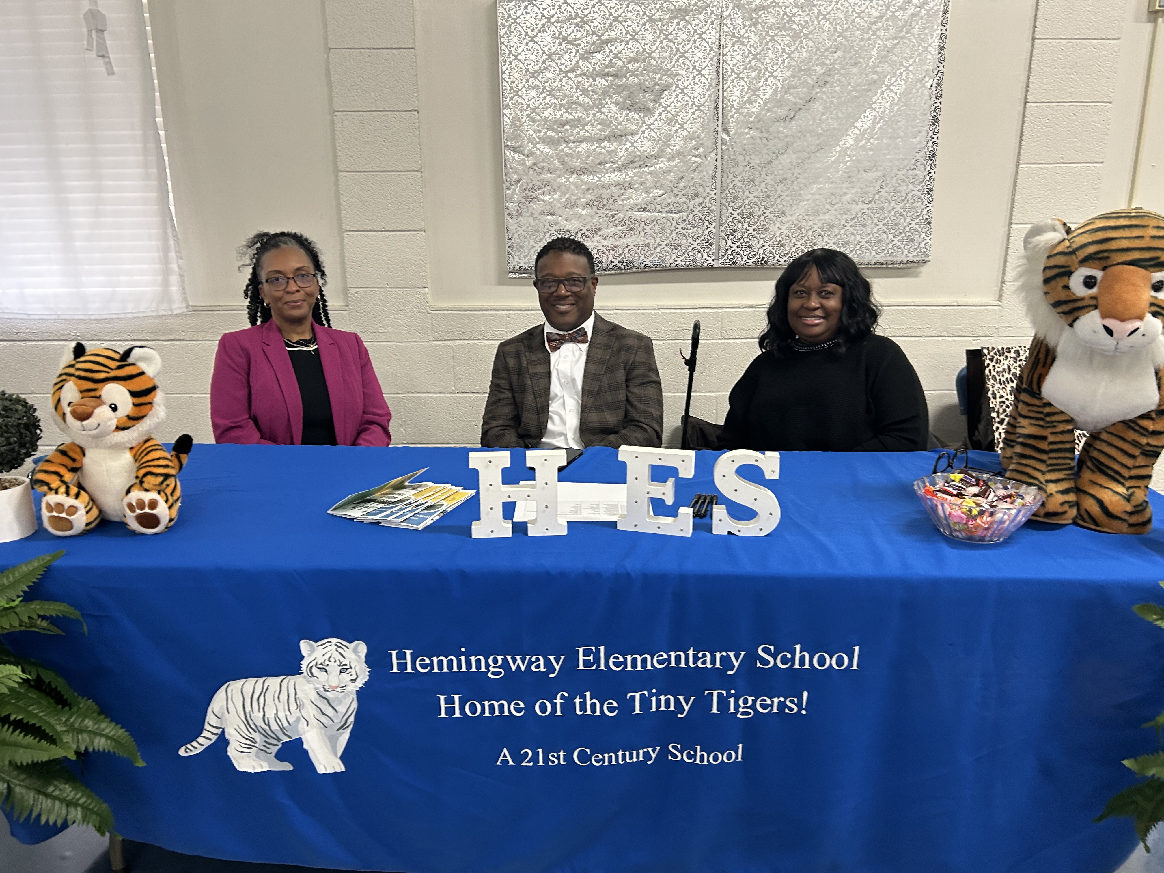 Four adults pose at a school information table inside a gym or multipurpose room. Three are seated behind a blue tablecloth that reads “Hemingway Elementary School – Home of the Tiny Tigers! A 21st Century School,” with white letters spelling “HES” on the table and a stuffed tiger mascot displayed beside brochures. A woman stands to the right of the table wearing glasses and a navy blazer. Decorative ferns frame the table, and bottled water and an exit sign are visible in the background.