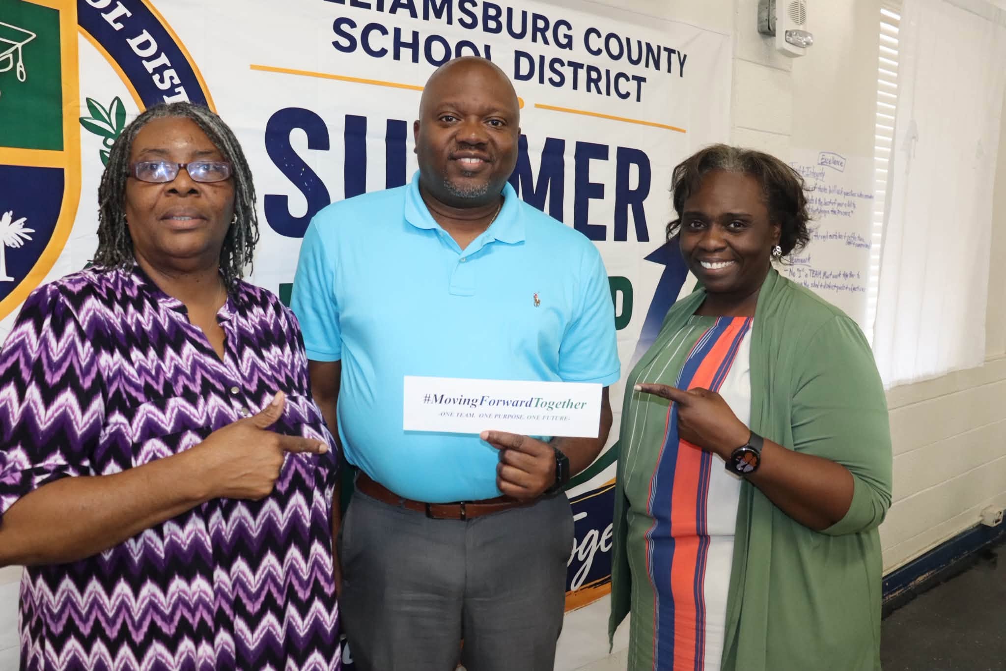 Three Williamsburg County School District staff members pose in front of a WCSD Summer Leadership backdrop. The person in the center holds a card reading “#MovingForwardTogether – One Team. One Purpose. One Future,” while the two staff members on either side smile and point toward the message.