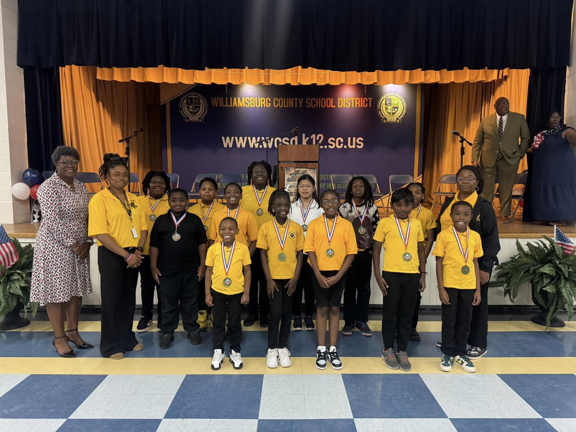 A group of elementary school students stand in front of a stage with a “Williamsburg County School District” banner and website displayed behind them. The students wear school uniforms or casual outfits and have medals around their necks, posing in a line. Several adults stand alongside them, one holding a trophy. The stage features gold curtains, chairs, microphones, and a podium, indicating an awards or recognition ceremony.