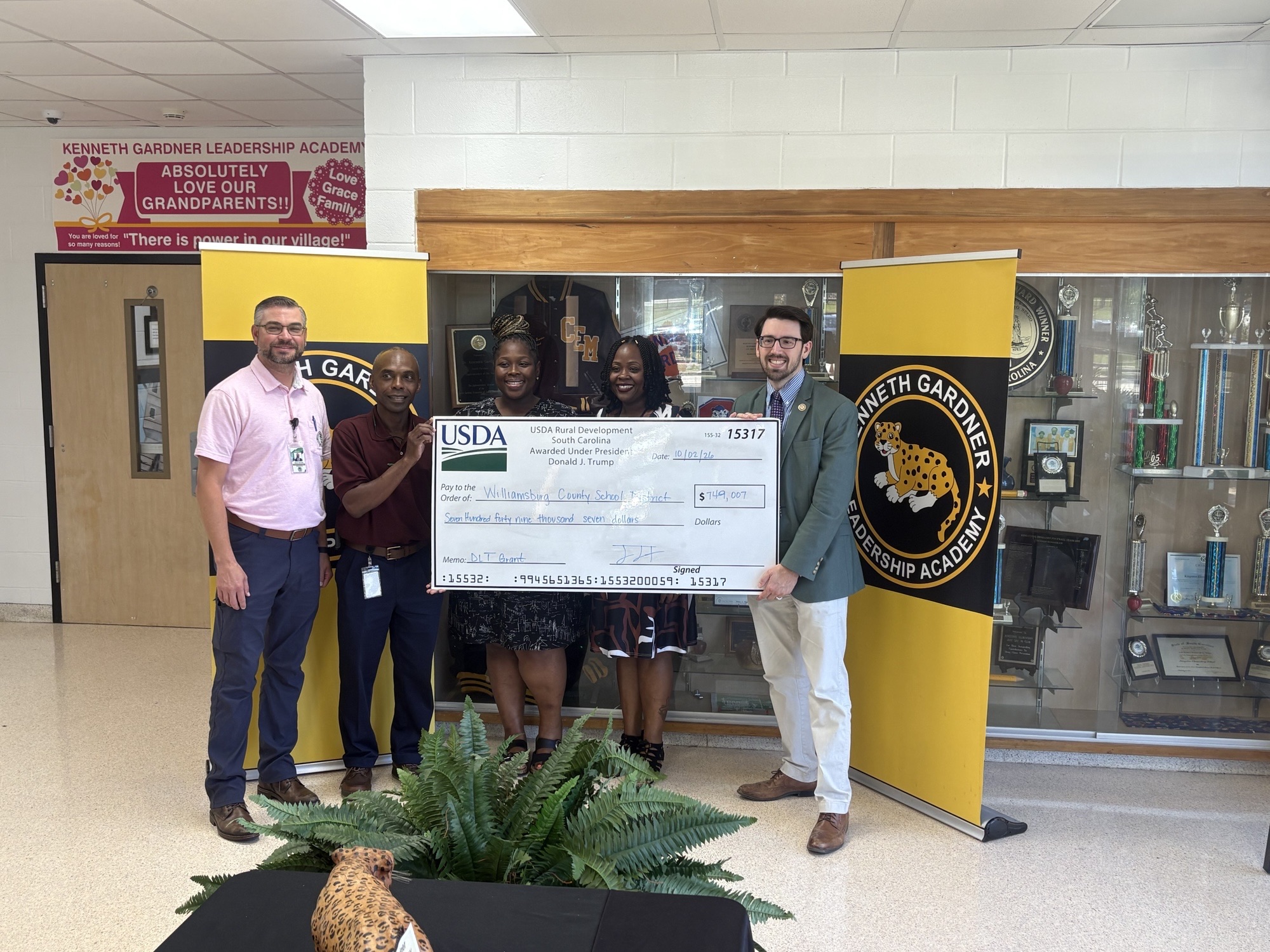 Five adults stand together in front of Kenneth Gardner Leadership Academy banners and a trophy display, holding a large USDA Rural Development South Carolina check for $749,007 made payable to Williamsburg County School District for a DLT grant. A potted fern and a spotted leopard statue sit in the foreground.