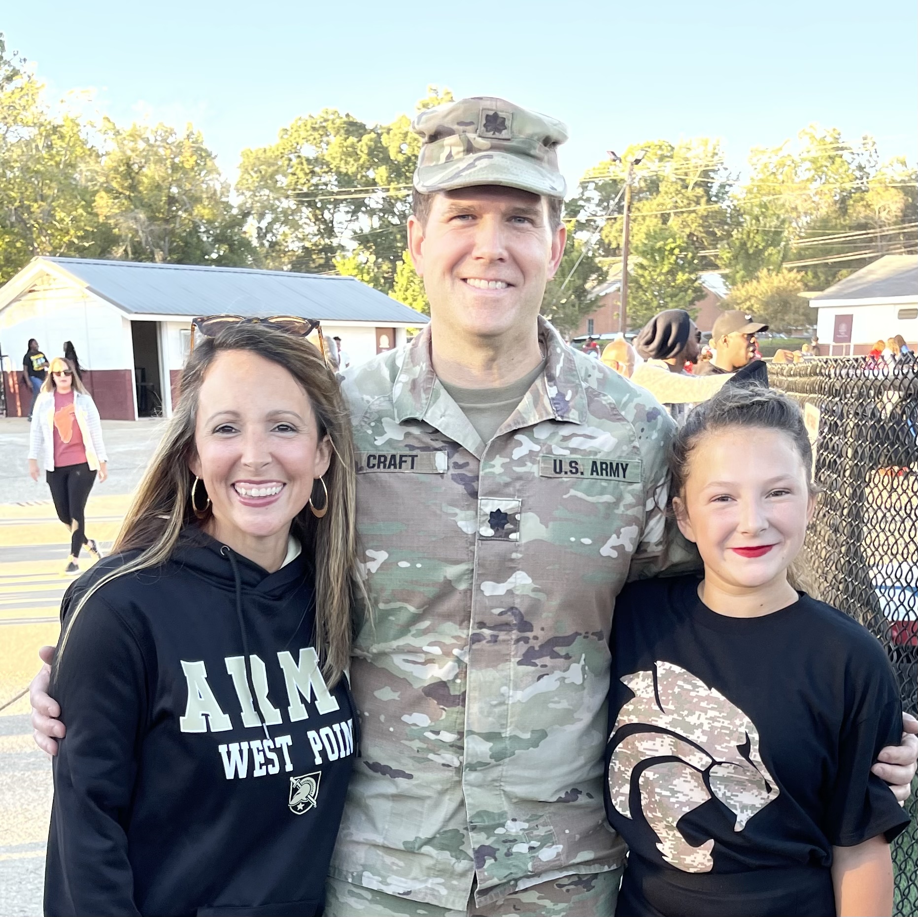Mrs. Craft standing to the left of her husband. Her husband is wearing military camo with their daughter,  standing to his right