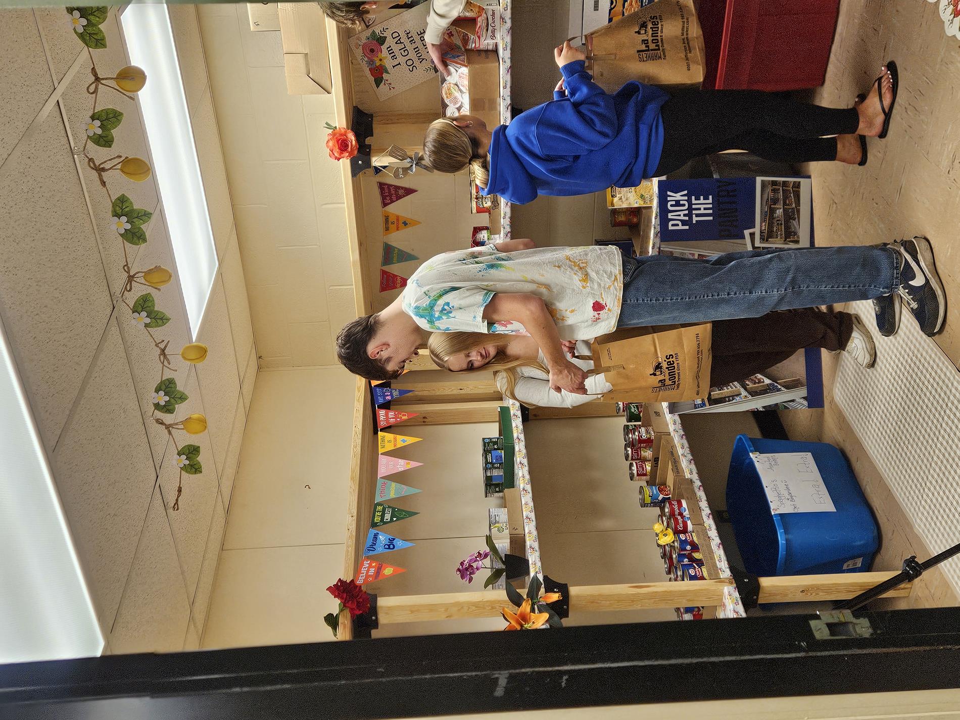 Two students pack non-perishable food items into a brown paper bag for a donation drive.