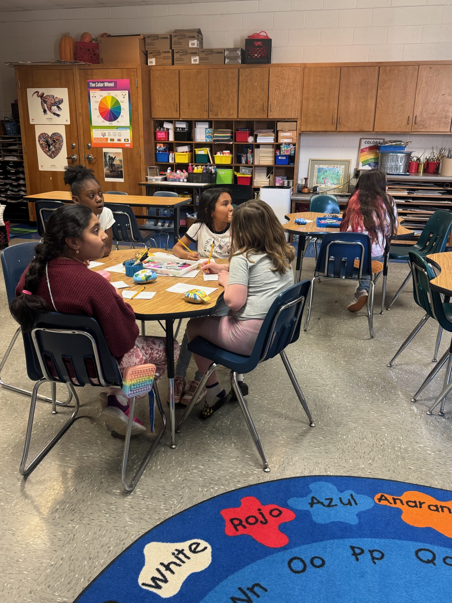 Students sit together at tables in an elementary art classroom, working on a colorful hands-on project. Art supplies, storage cabinets, posters, and a bright color-themed rug fill the classroom.