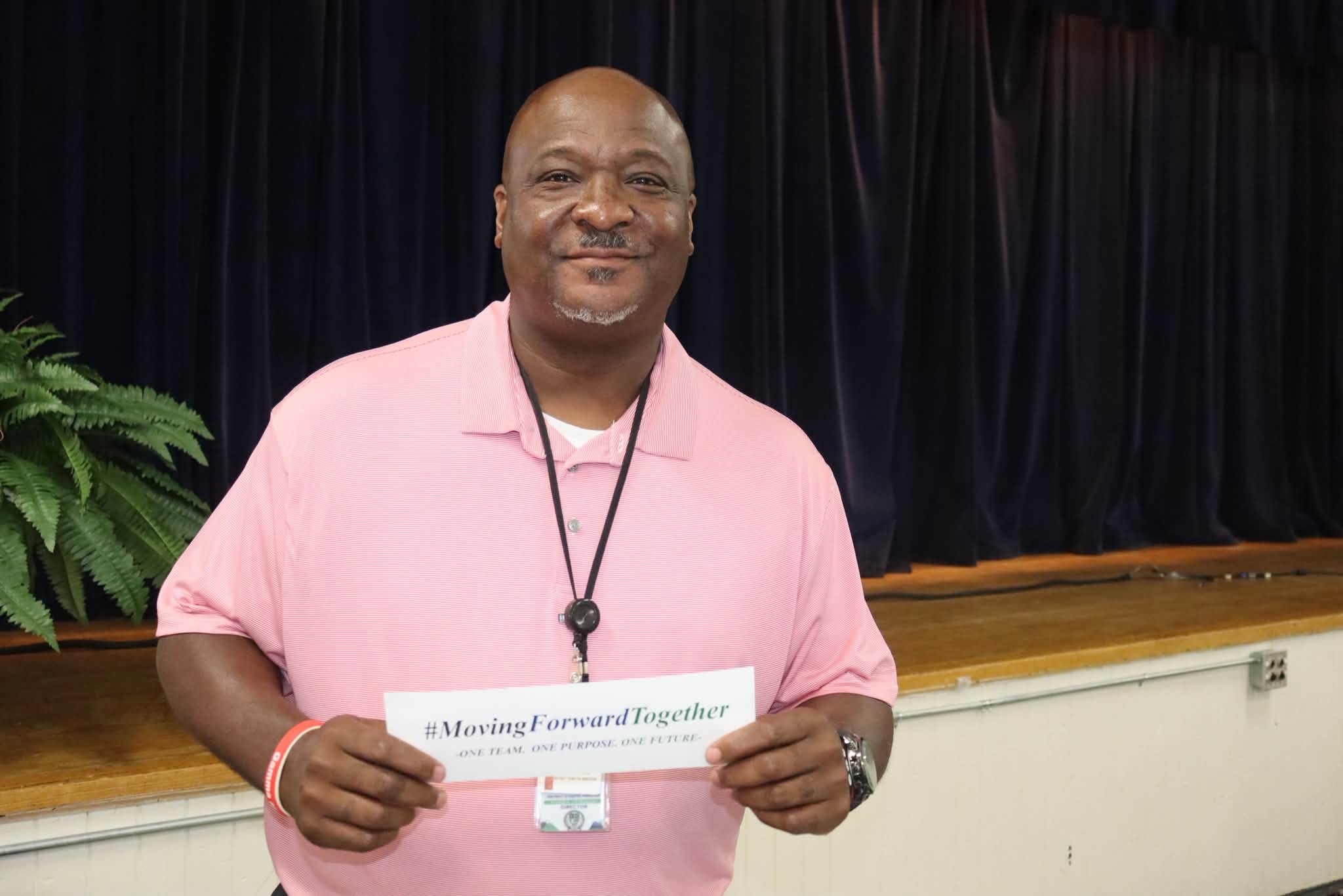 A smiling Williamsburg County School District staff member wearing a light pink polo shirt stands in front of a stage with dark curtains, holding a card that reads “#MovingForwardTogether – One Team. One Purpose. One Future.” while wearing a district ID badge on a lanyard.