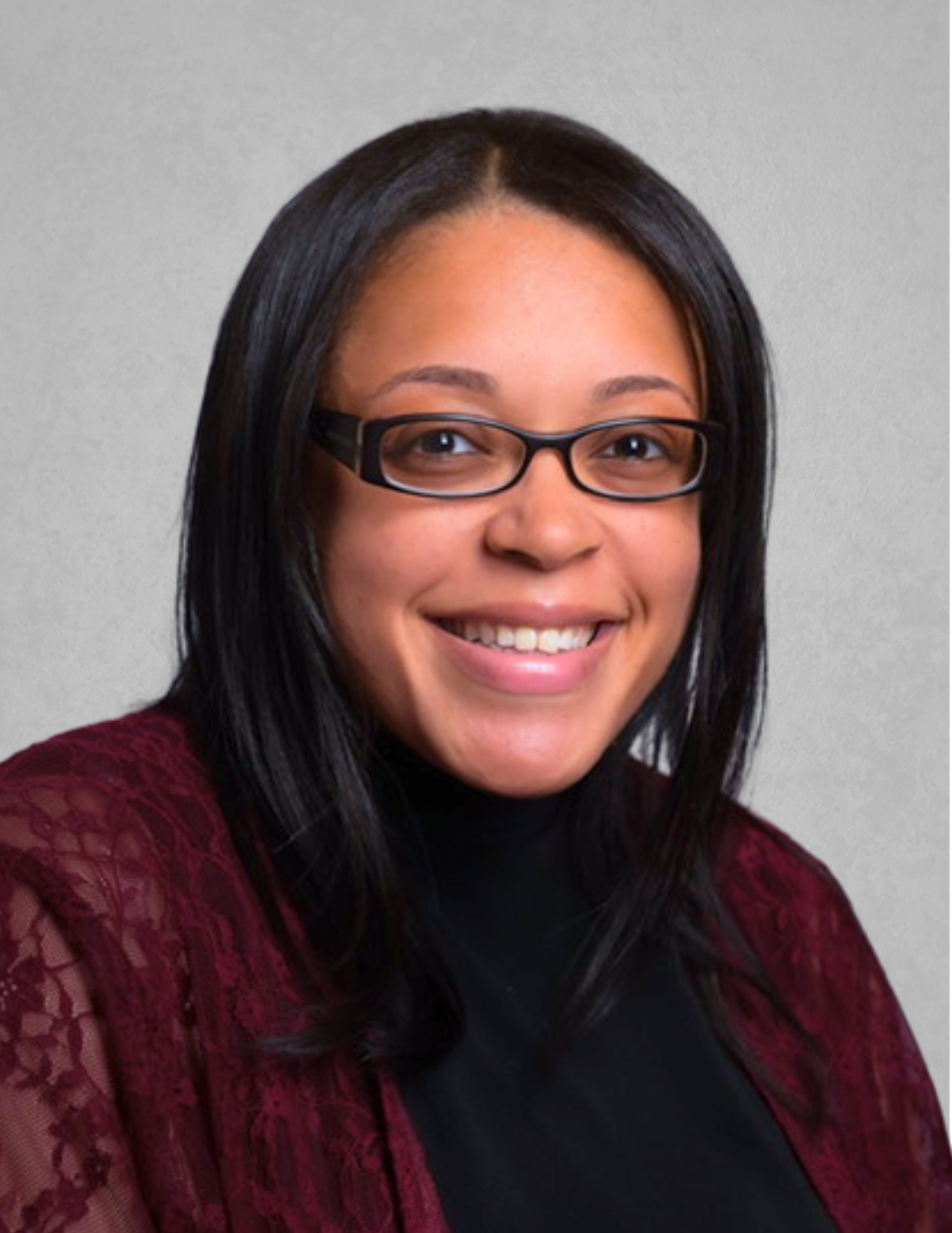 Professional headshot of Alisha Williams smiling and facing the camera. She is wearing black-framed glasses, a black top, and a burgundy lace cardigan. Williams has straight dark hair worn past her shoulders and is posed against a light gray background.