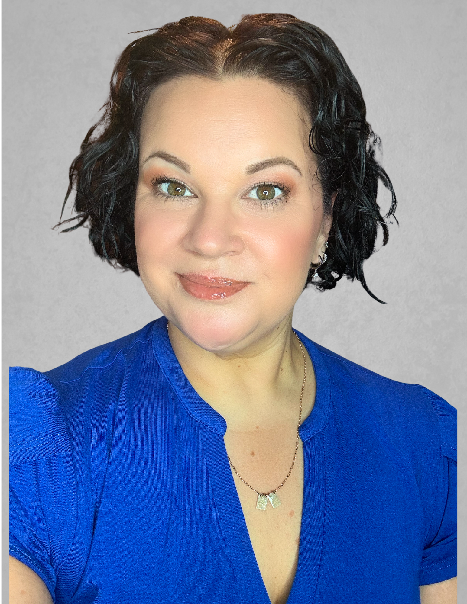 Professional headshot of Sarah Dupont smiling and facing the camera. She is wearing a royal blue blouse and a silver necklace, with short dark curly hair and light makeup. The portrait is set against a light gray background.