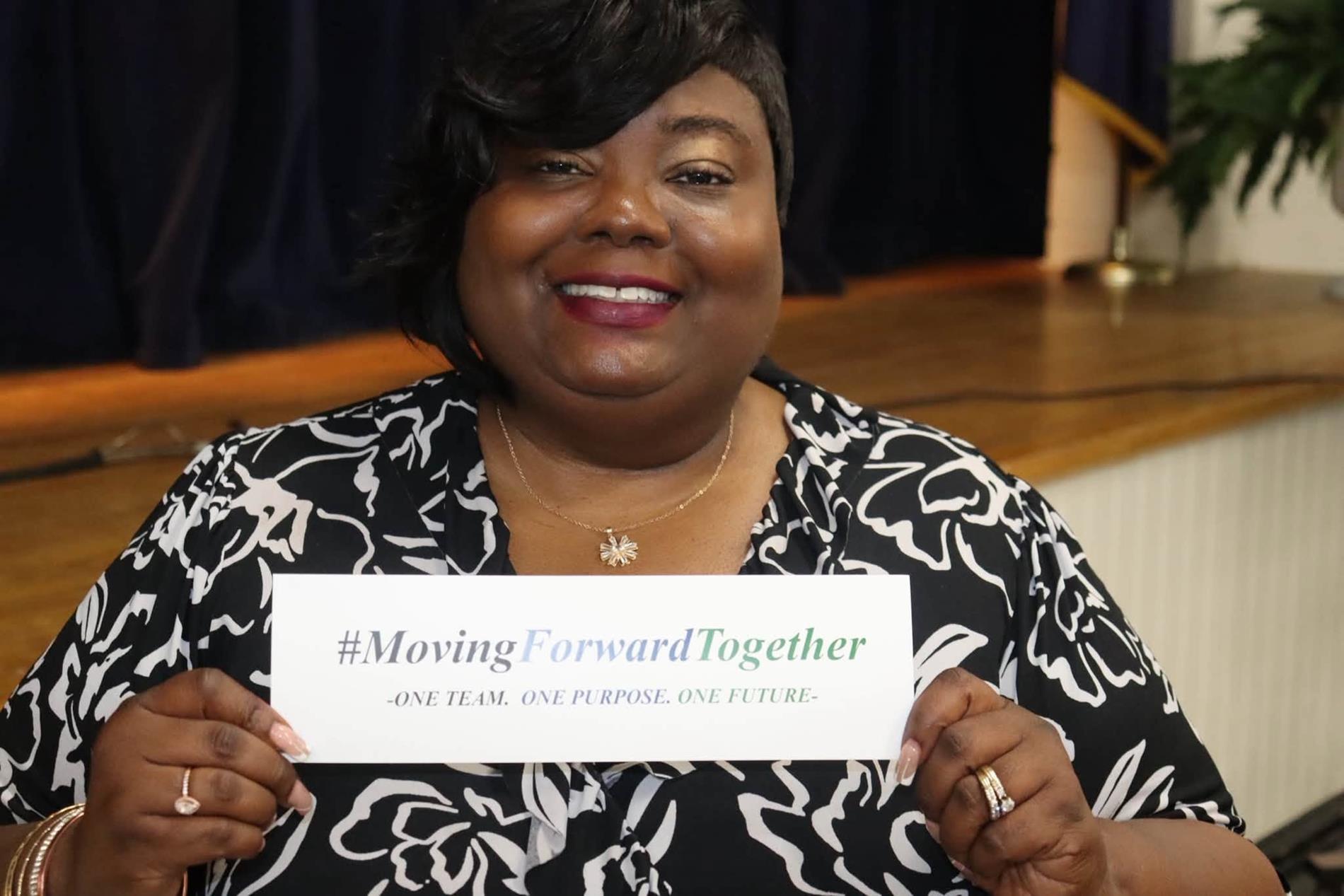  A smiling Williamsburg County School District staff member wearing a black-and-white floral blouse holds a card reading “#MovingForwardTogether – One Team. One Purpose. One Future.” She is seated in front of a stage with dark curtains during a Summer Leadership event.