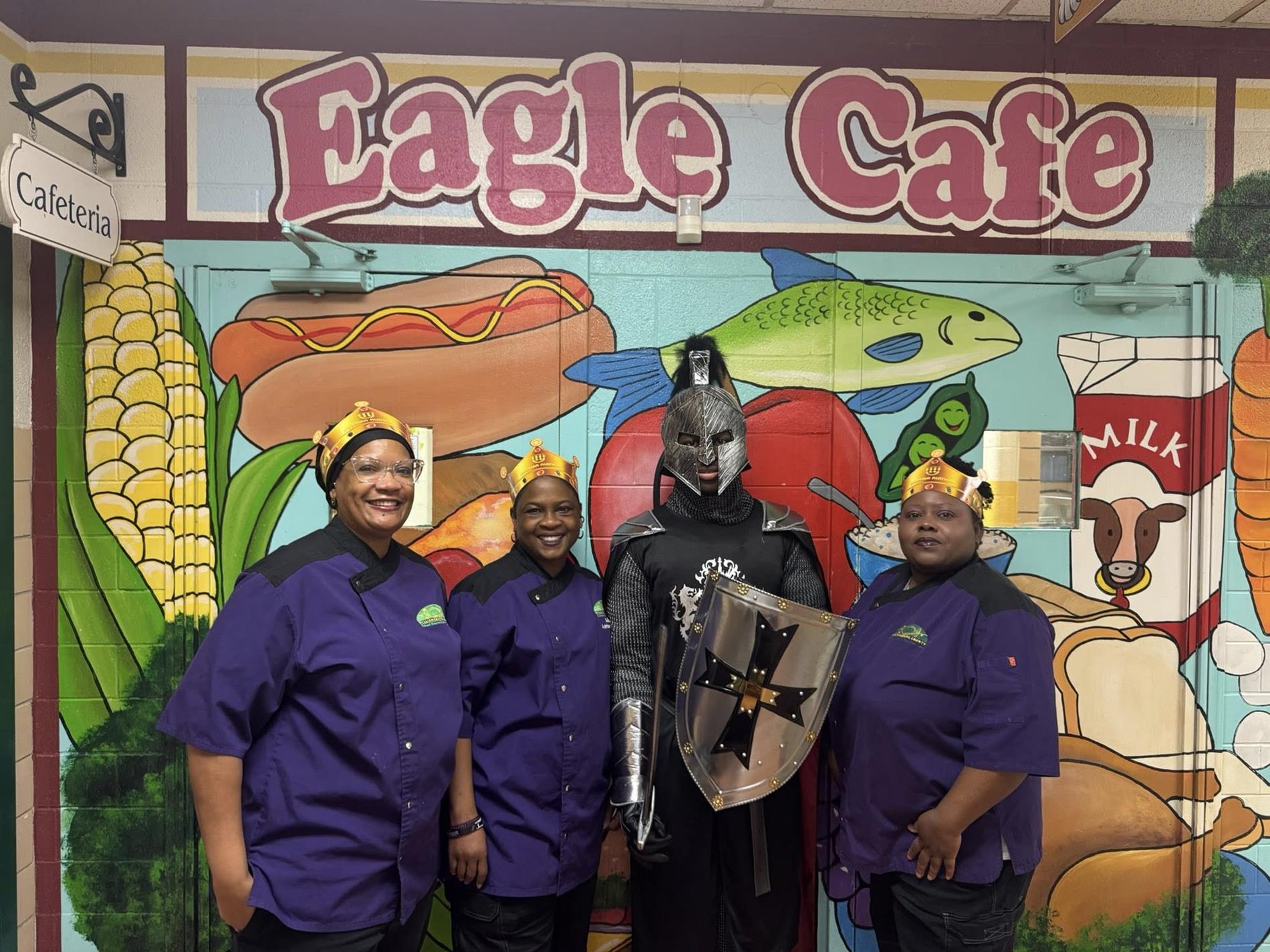 Four adults pose in front of a colorful “Eagle Cafe” mural inside a cafeteria. Three women wearing matching purple work uniforms and gold paper crowns stand smiling beside a person dressed as a medieval knight in black and silver armor, holding a shield with a black cross emblem. The mural behind them features large illustrations of food items including corn, a hot dog, a fish, apples, milk, and bread, along with a hanging “Cafeteria” sign.