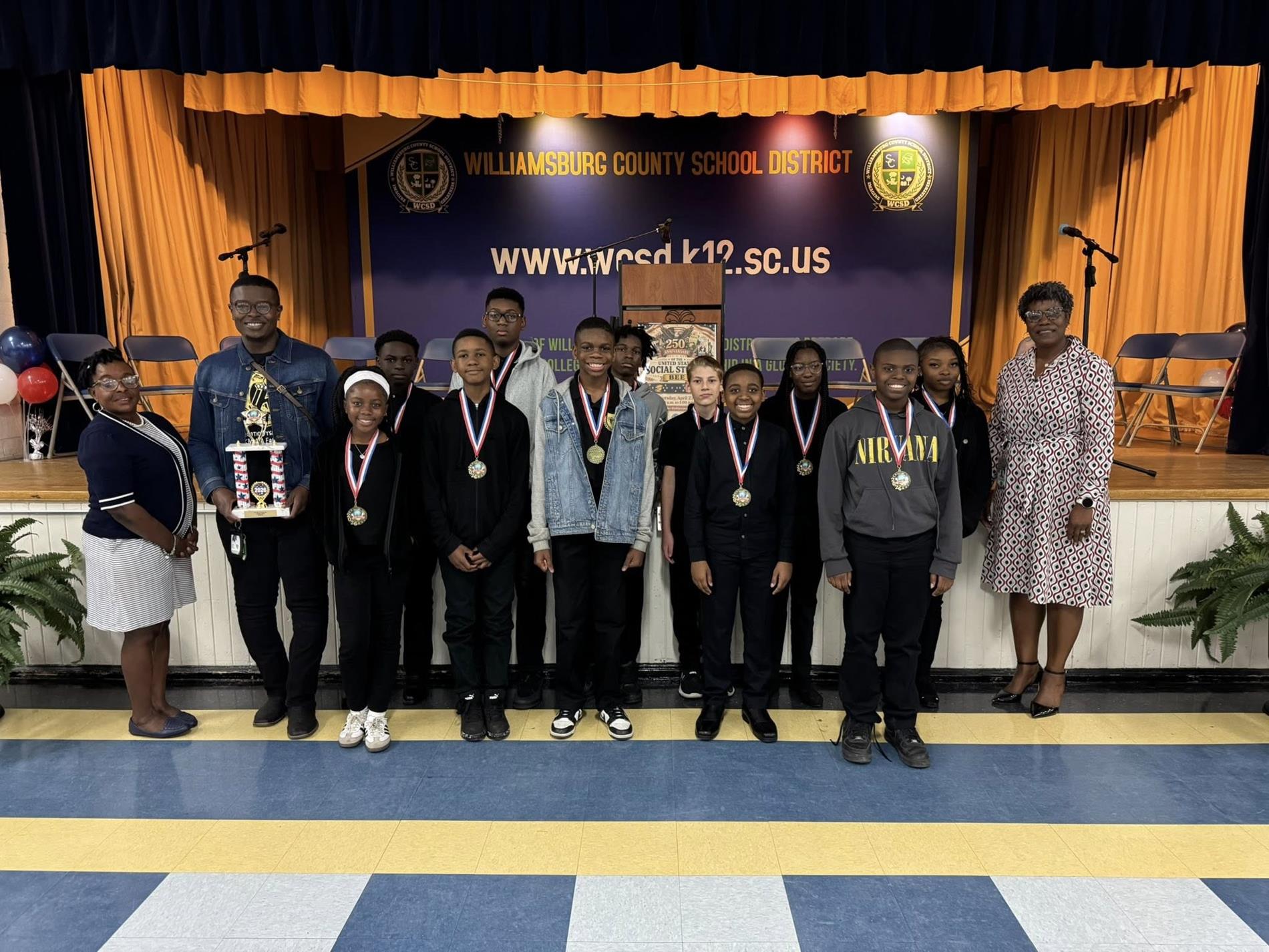 A group of middle school students stand in front of a stage with a “Williamsburg County School District” banner and website displayed behind them. The students wear school uniforms or casual outfits and have medals around their necks, posing in a line. Several adults stand alongside them, one holding a trophy. The stage features gold curtains, chairs, microphones, and a podium, indicating an awards or recognition ceremony.