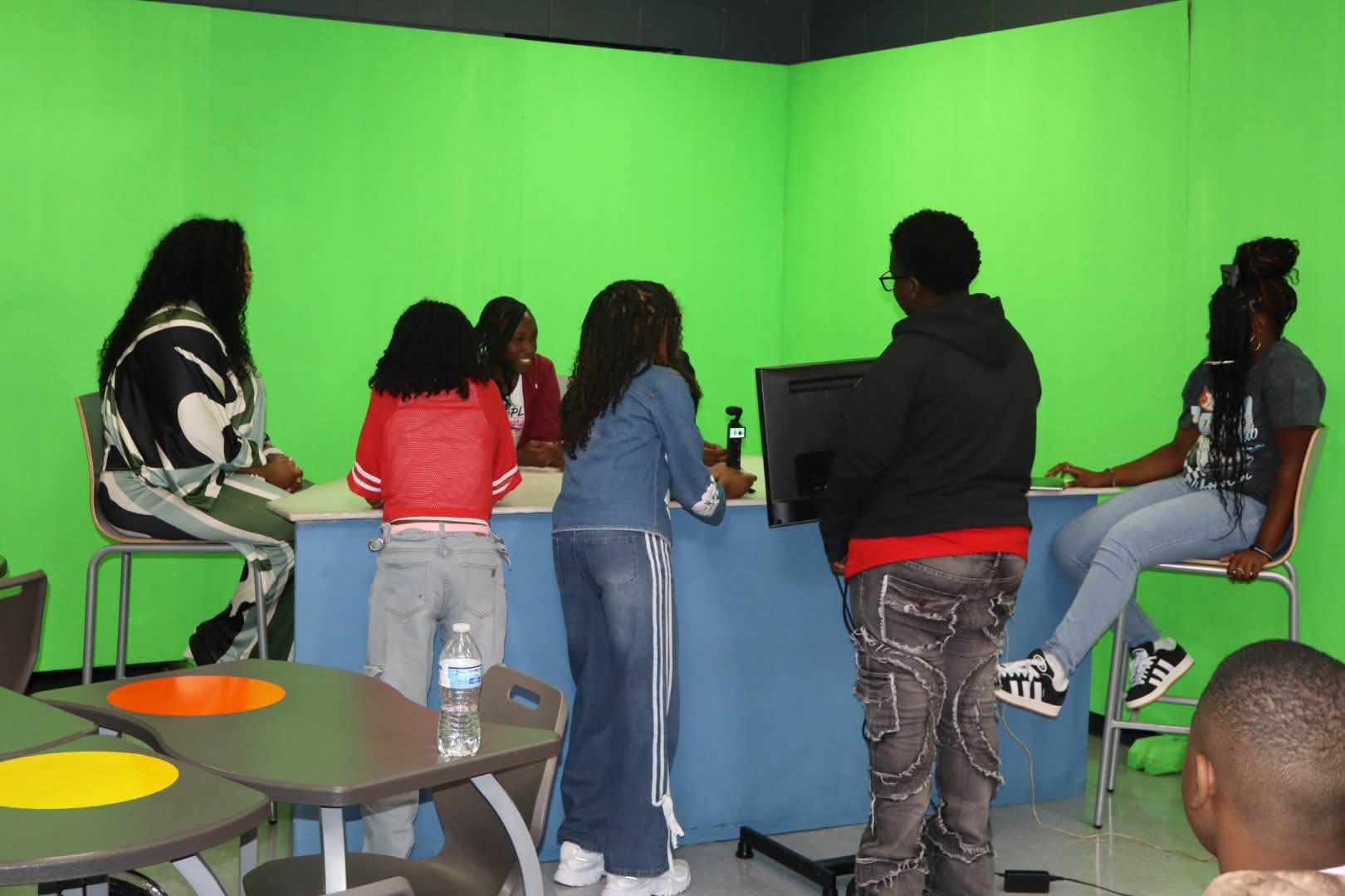 A group of students and an adult gather around a desk in a green-screen studio. Several students sit on stools or stand near a computer monitor and microphone, appearing to participate in a media or video production activity.