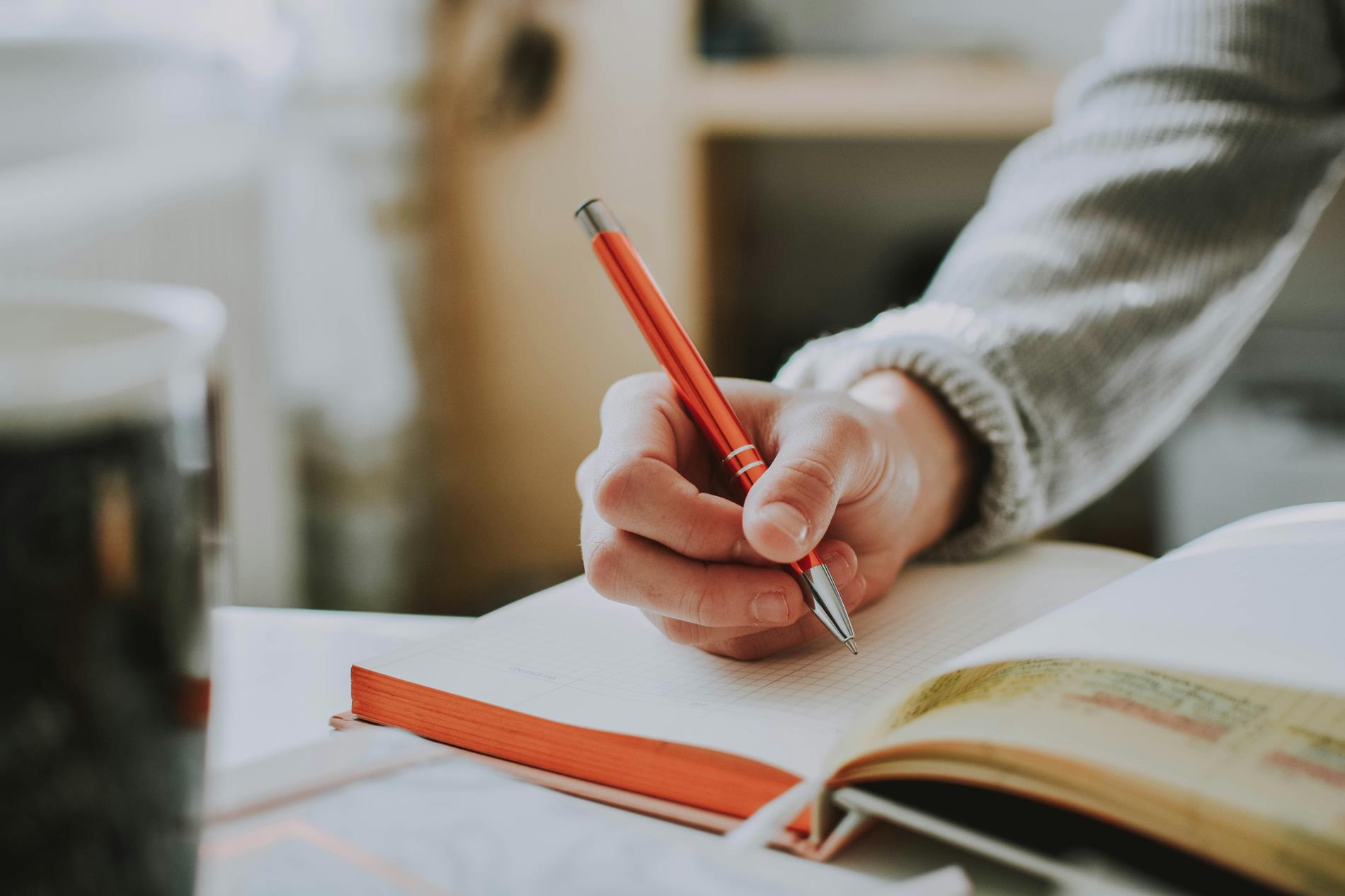 A woman's hand holds a red pen and writes in a notebook on a desk