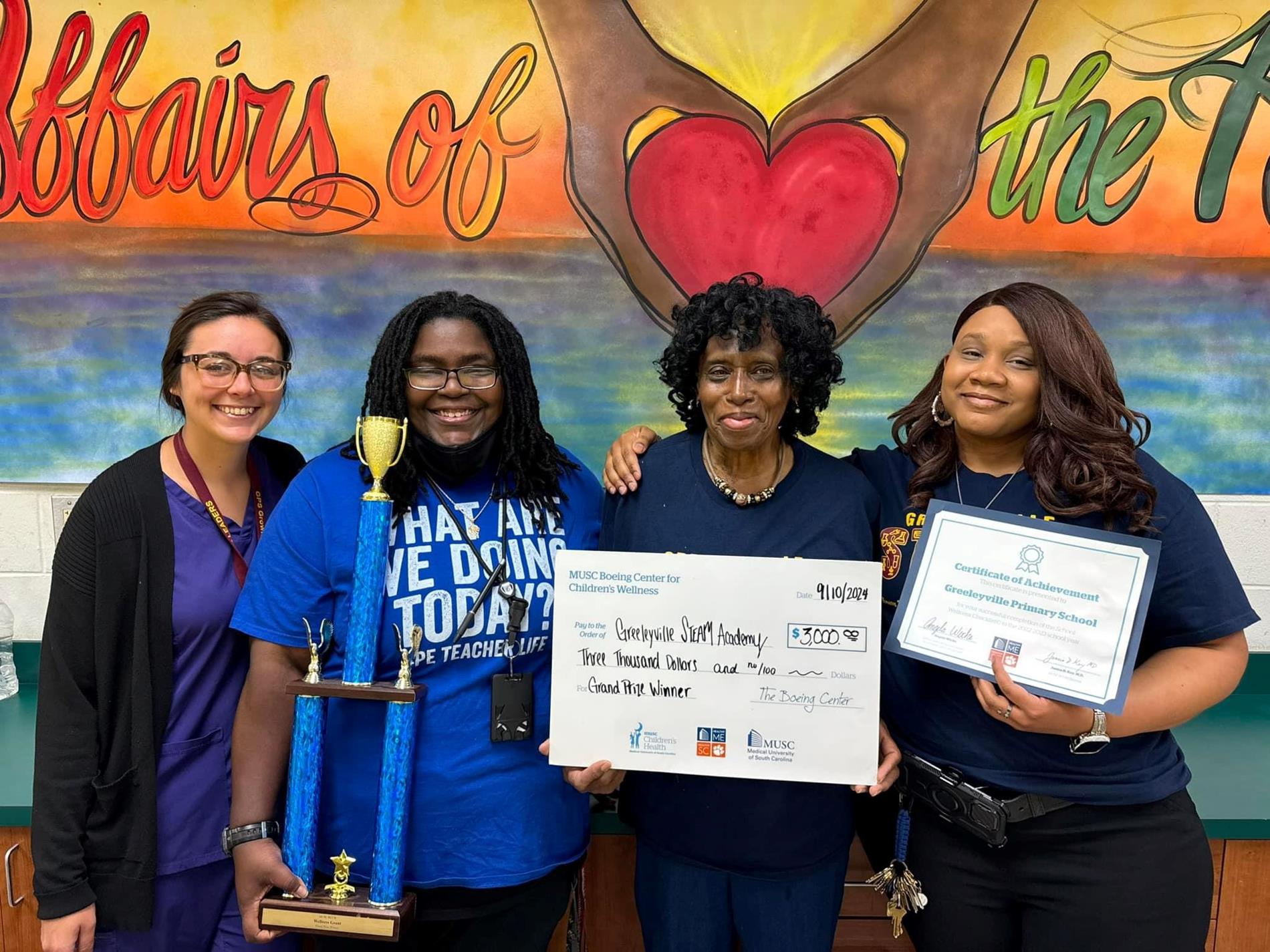 picture of four ladies. one is holding a trophy. one is holding a check. one is holding an award certificate.