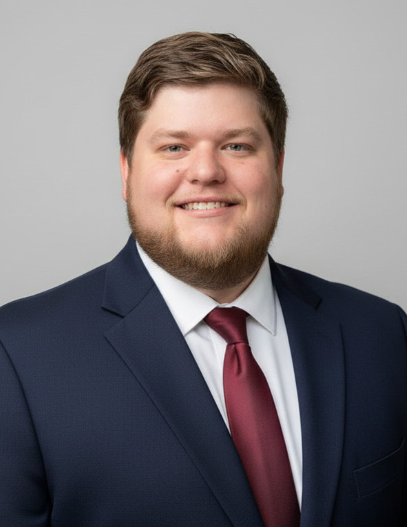 Professional headshot of Corey Alleman wearing a navy suit, white dress shirt, and burgundy tie. He is smiling and facing the camera against a light gray background. Alleman has short brown hair and a neatly groomed beard.