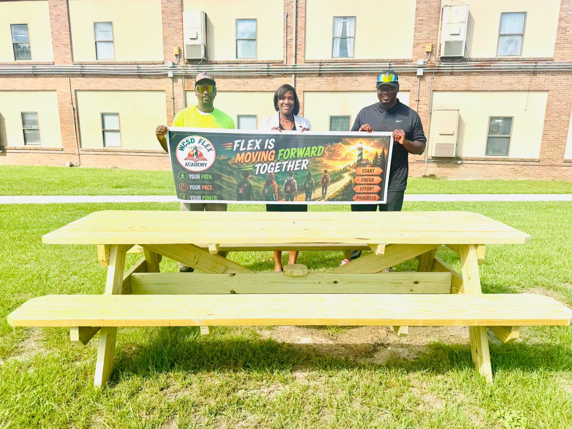 Three people stand outdoors on a grassy lawn in front of a brick school building, holding a WCSD FLEX Academy banner that reads, “FLEX Is Moving Forward Together.” The banner also highlights “Your Path. Your Pace. Your Power.” and features a pathway graphic with signs reading “Start, Focus, Effort, Progress.” A newly constructed wooden picnic table is positioned in the foreground.