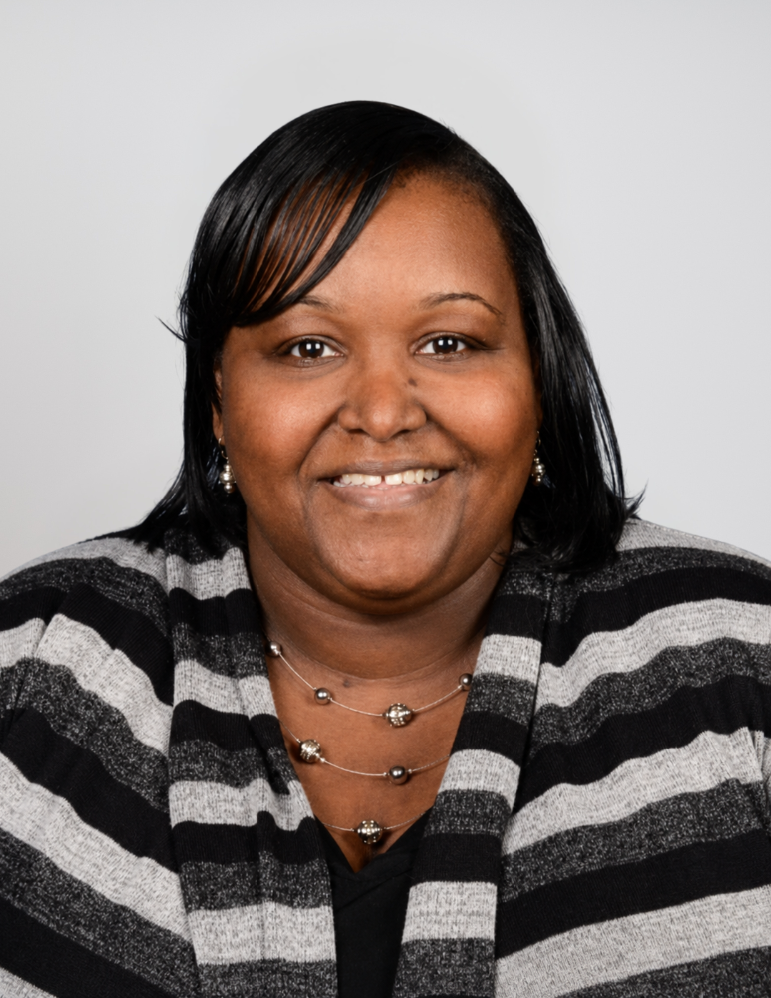 Professional headshot of Caneshia Jacob smiling and facing the camera. She is wearing a black and gray striped sweater with layered silver necklaces and small earrings. Her shoulder-length black hair is styled with a side-swept fringe, and she is posed against a light gray background. The portrait is suitable for use in an instructional coach or school leadership announcement.