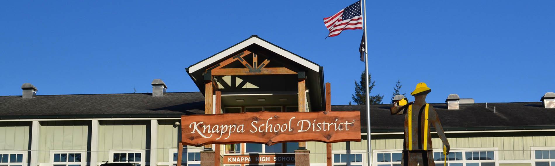 Knappa School District sign and metal Logger in front of the high school building with American flag in the background