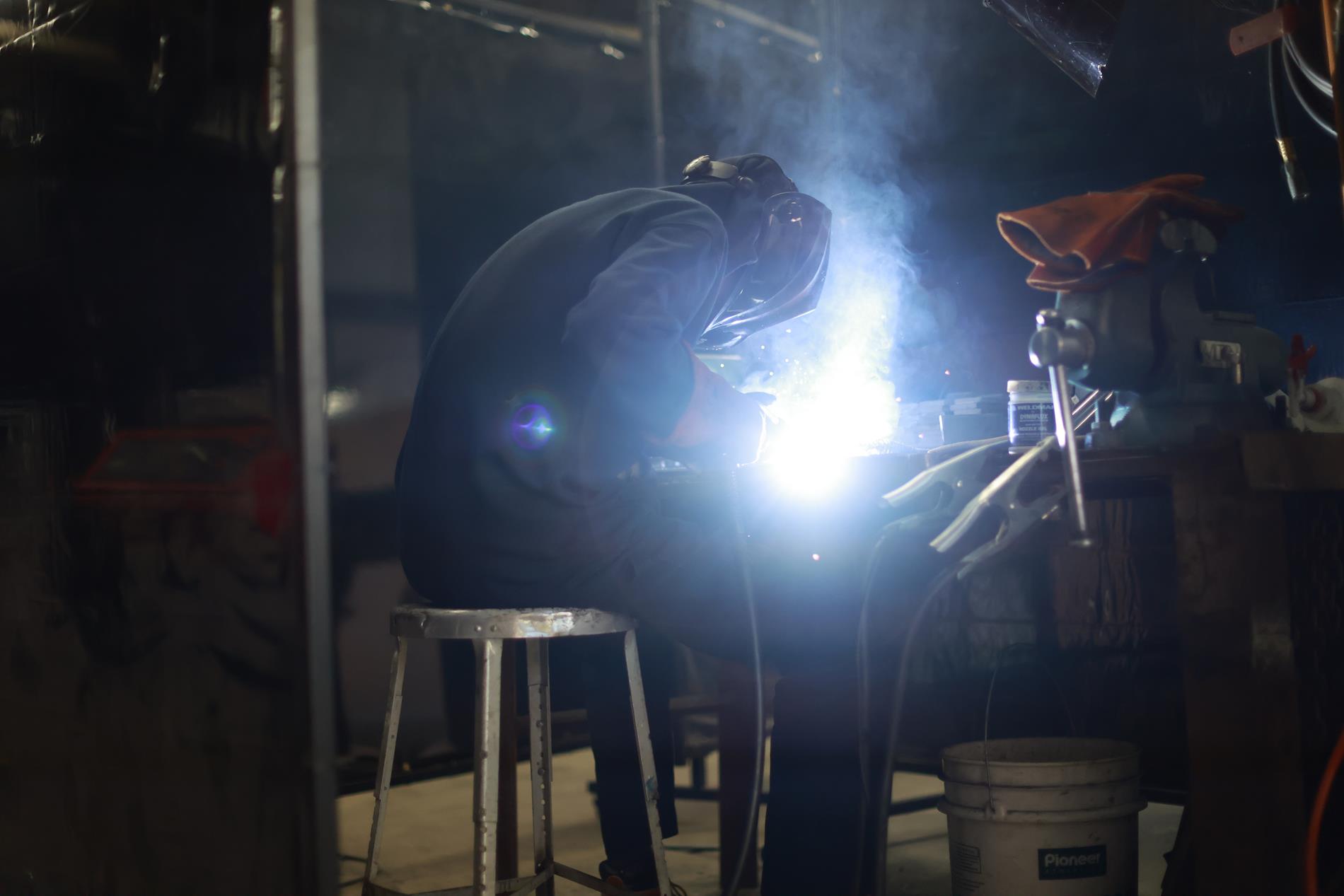 A high school student in a welding helmet and suit welds a piece of metal 