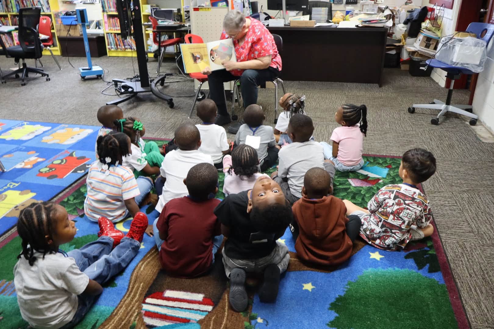 A teacher sits at the front of a classroom or school library reading a picture book to a group of young students seated on a colorful rug. The children face the teacher and book, while bookshelves, desks, chairs, and classroom supplies fill the background.