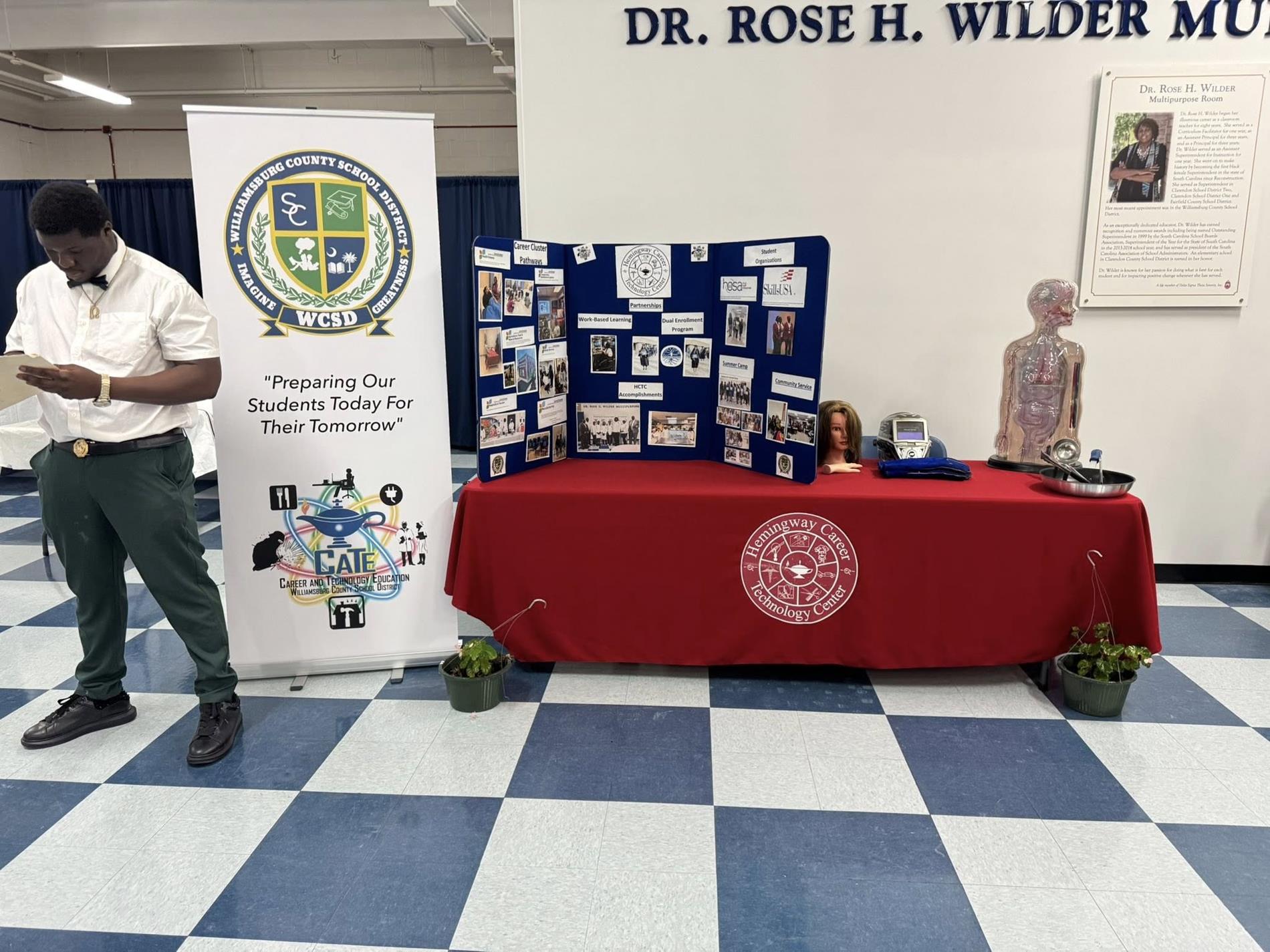 A student in a white shirt and green pants stands looking at his phone beside an informational display table inside a school building. The table, covered with a red “Hemingway Career & Technology Center” tablecloth, features a blue tri-fold board with photos and program information, a CPR training mannequin torso, a mannequin head, medical equipment, and a metal tray. A standing banner for the Williamsburg County School District reads “Preparing Our Students Today For Their Tomorrow.” The floor has blue-and-white checkered tiles, and a wall sign above reads “Dr. Rose H. Wilder.”