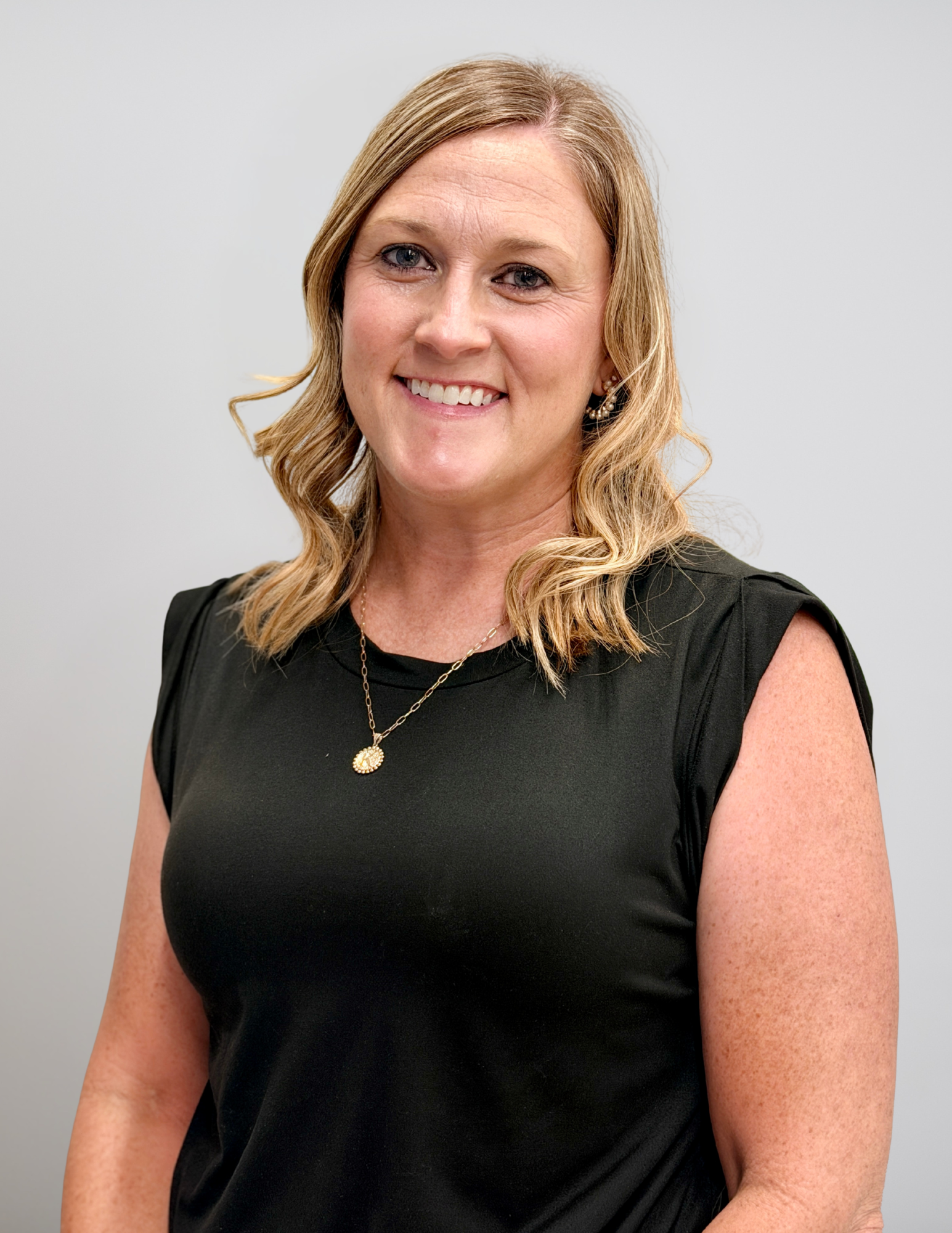 Professional headshot of Kelly Juneau smiling and facing the camera. She is wearing a black sleeveless blouse, a gold pendant necklace, and small earrings. Her shoulder-length blonde hair is styled in loose waves. She is standing against a light gray background with a friendly and approachable expression. The portrait is suitable for use in an instructional coach or school leadership announcement.