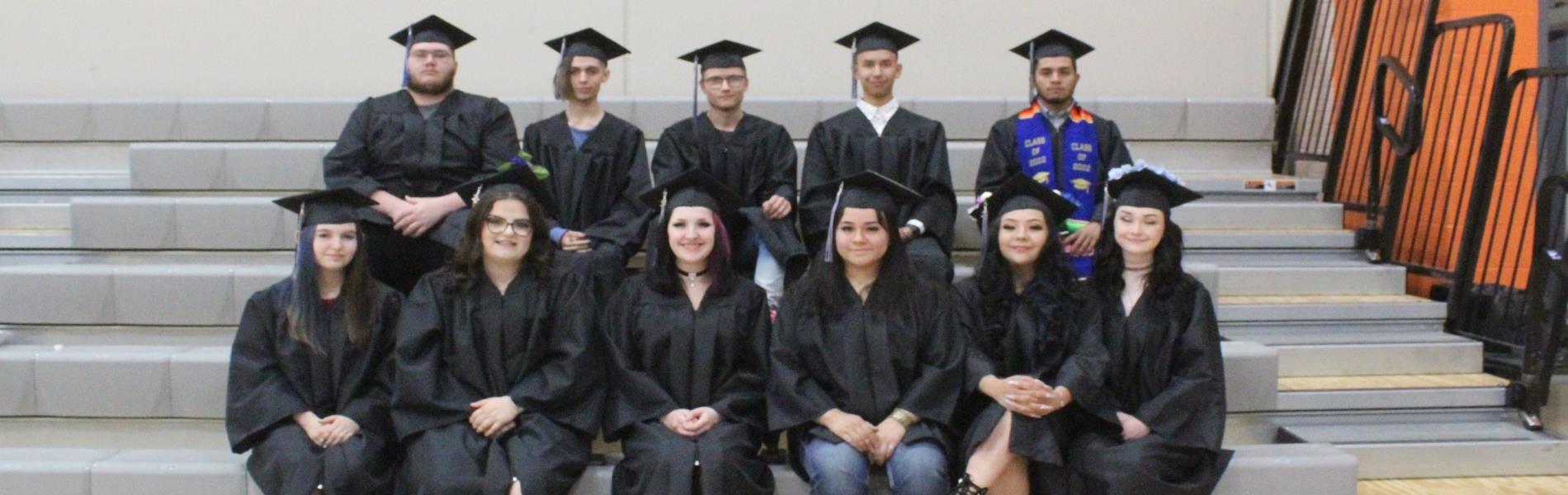 Eleven graduates dressed in black caps and gowns sit in two rows on gray bleachers. 