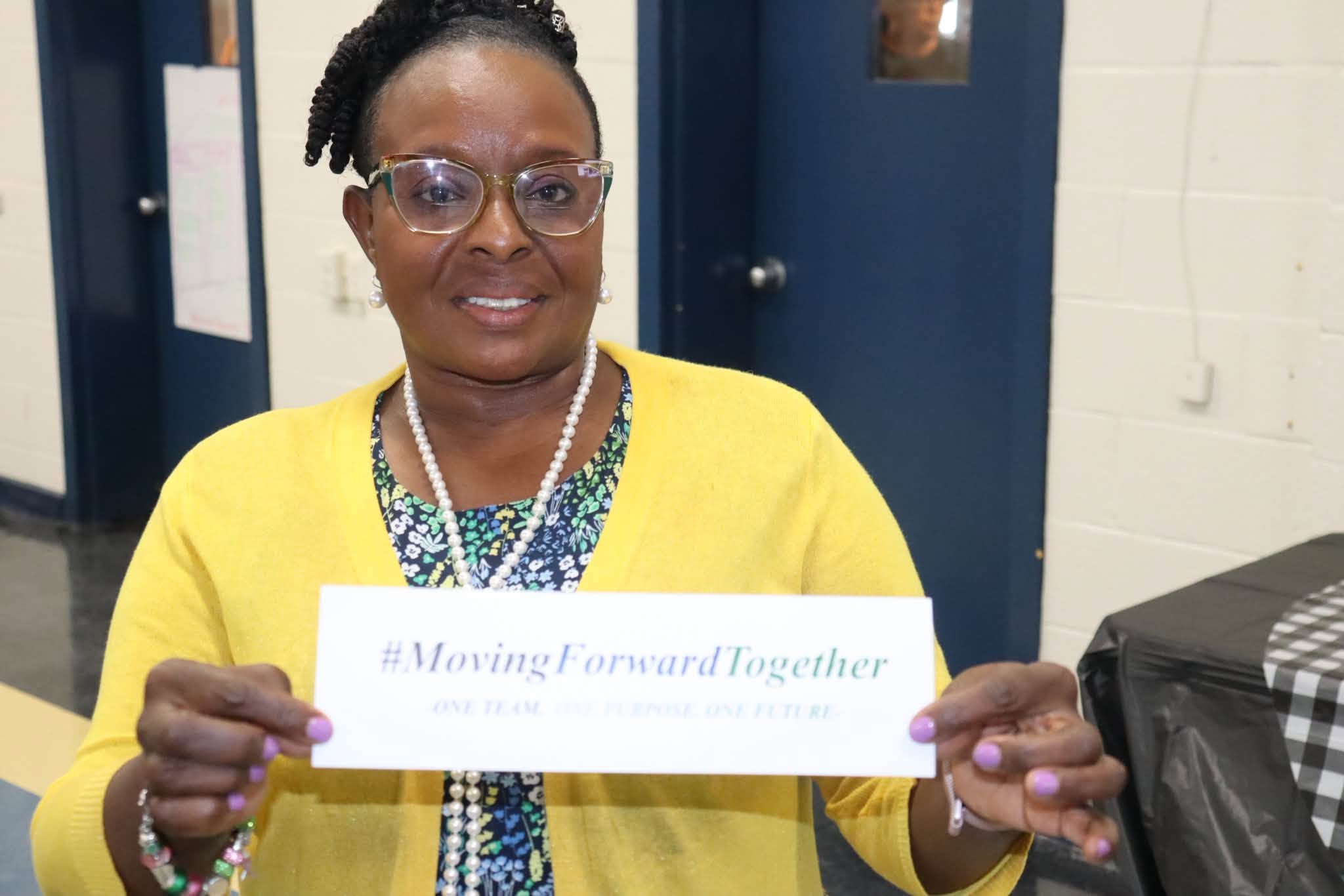 A smiling Williamsburg County School District staff member wearing glasses, a yellow cardigan, and a pearl necklace holds a card reading “#MovingForwardTogether – One Team. One Purpose. One Future.” while seated at a table during a Summer Leadership event.
