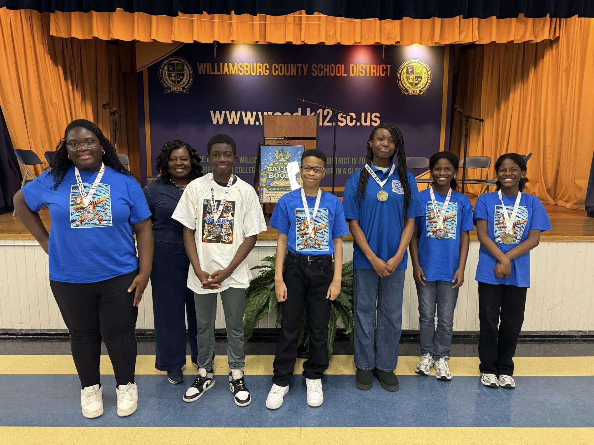 students standing in front of a blue and orange curtained backdrop with a banner that reads "WILLIAMSBURG COUNTY SCHOOL DISTRICT" and the district's website, "www.wcsd.k12.sc.us." 