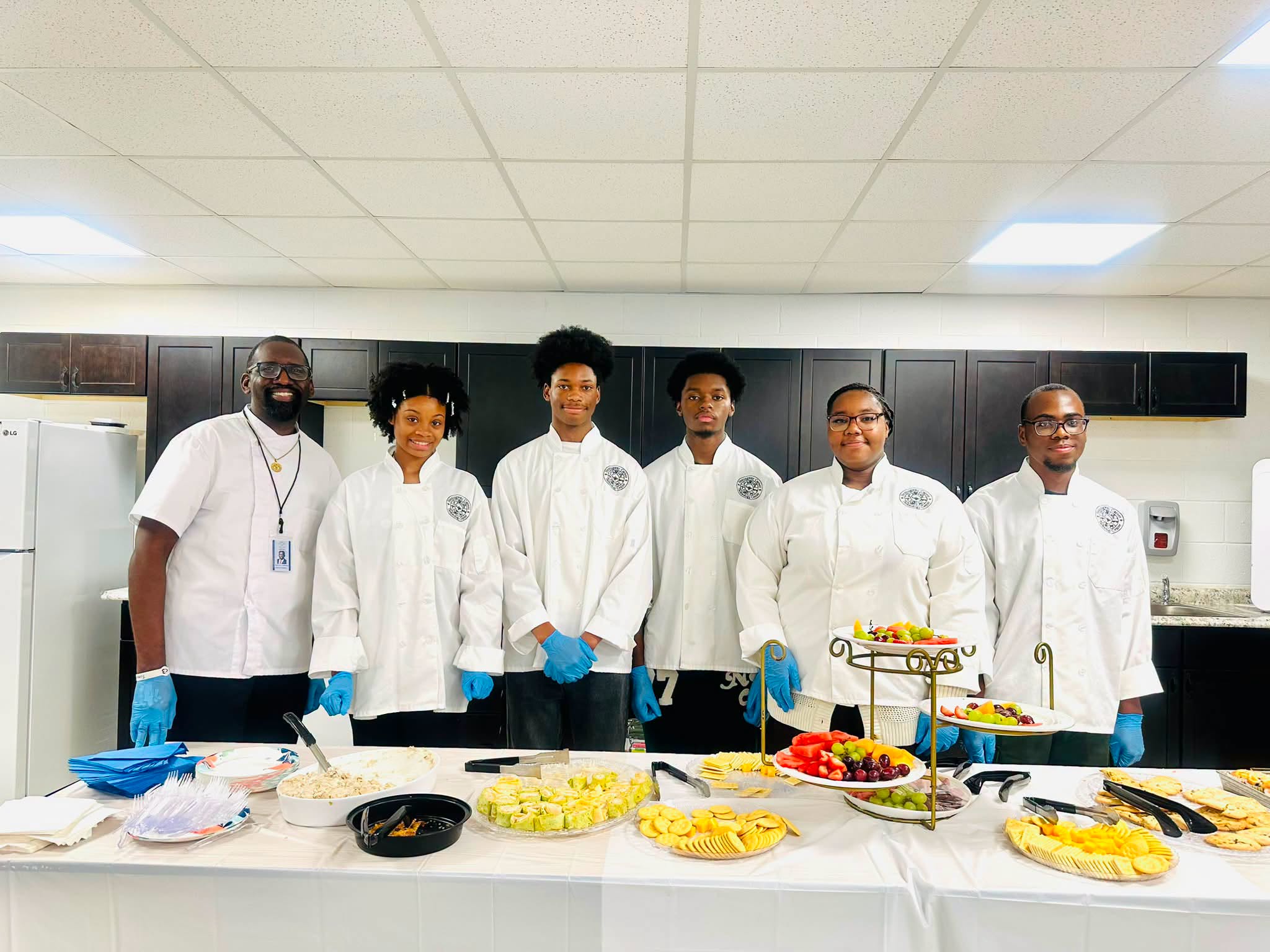 Six people wearing white chef coats stand together in a culinary classroom behind a table filled with prepared foods, including fruit, vegetables, crackers, cheese, and other refreshments. Several members of the group are wearing blue food-service gloves, and dark kitchen cabinets and appliances are visible in the background.