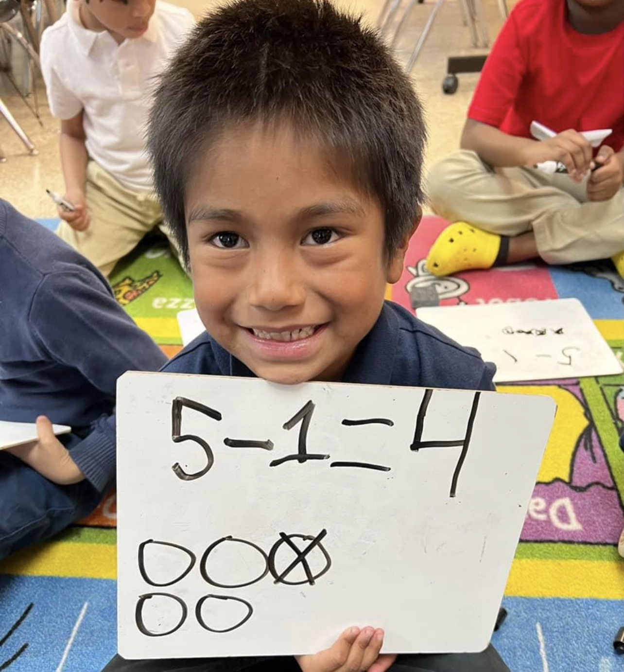 Kindergarten student smiling showing his math equation 5-4=1. He drew a picture on a white board showing how he solved the problem.