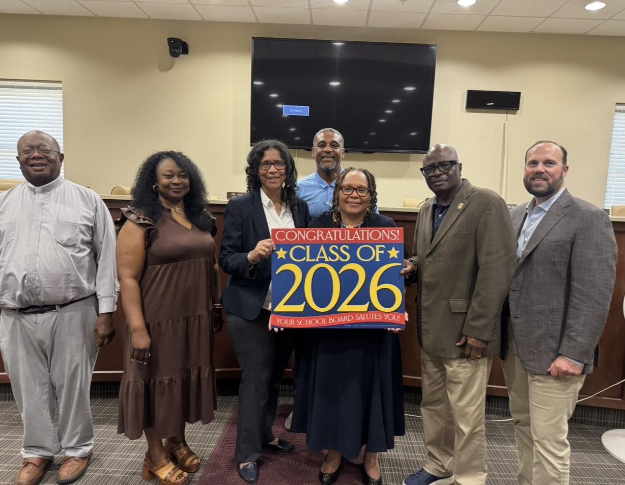 A group of six adults stands together in a school board or meeting room, smiling for a photo. Two women in the center hold a colorful sign that reads, “Congratulations! Class of 2026 — Your School Board Salutes You!” The group includes men and women dressed in business and casual attire, standing in front of a large wall-mounted monitor and conference-style seating. The room is brightly lit with ceiling lights and has windows with blinds along the walls.