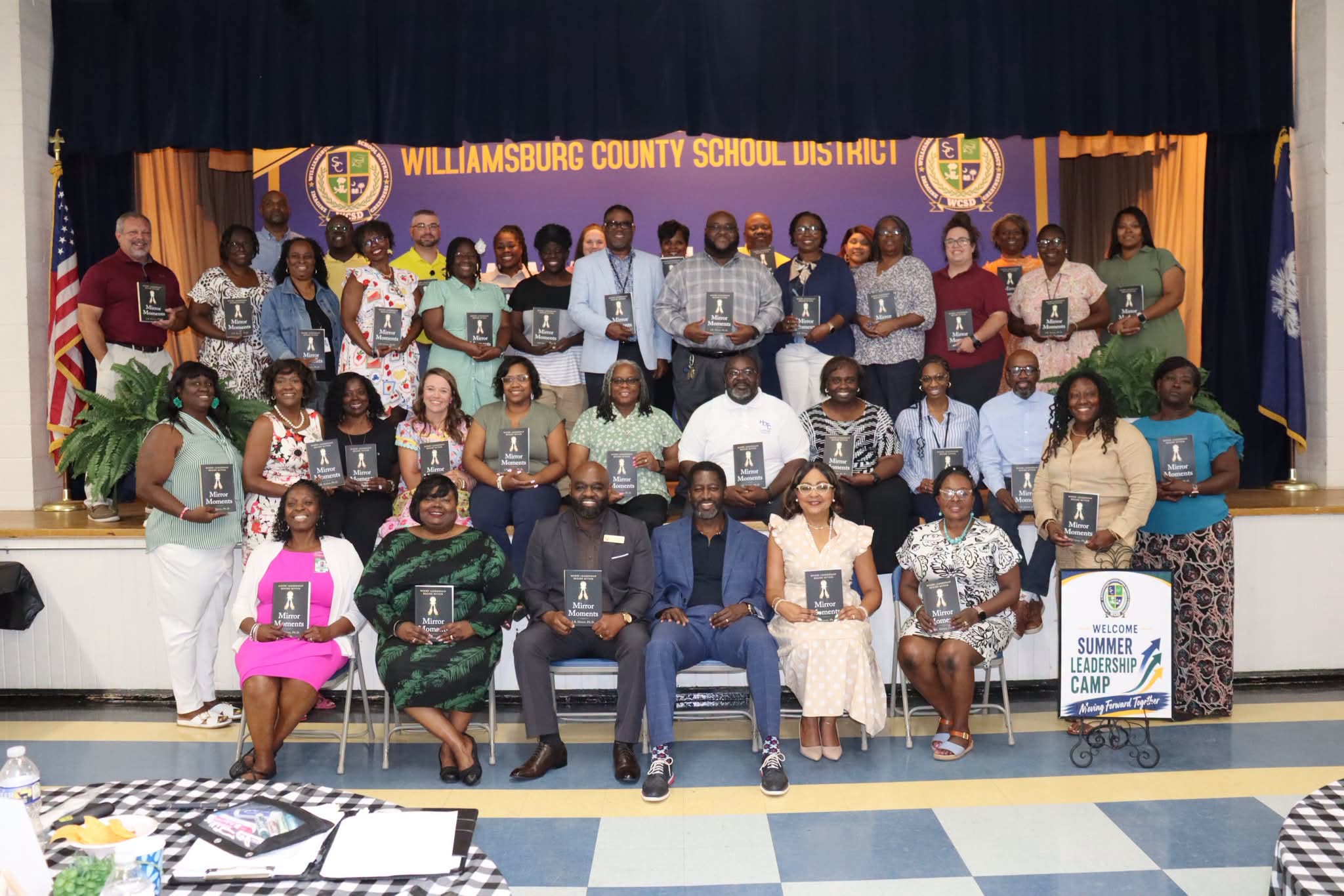 A large group of Williamsburg County School District educators and staff pose together on a stage during Summer Leadership Camp, each holding a copy of the book *Mirror Moments*. A Summer Leadership Camp welcome sign is displayed at the front of the stage, with WCSD branding and the district banner visible in the background.