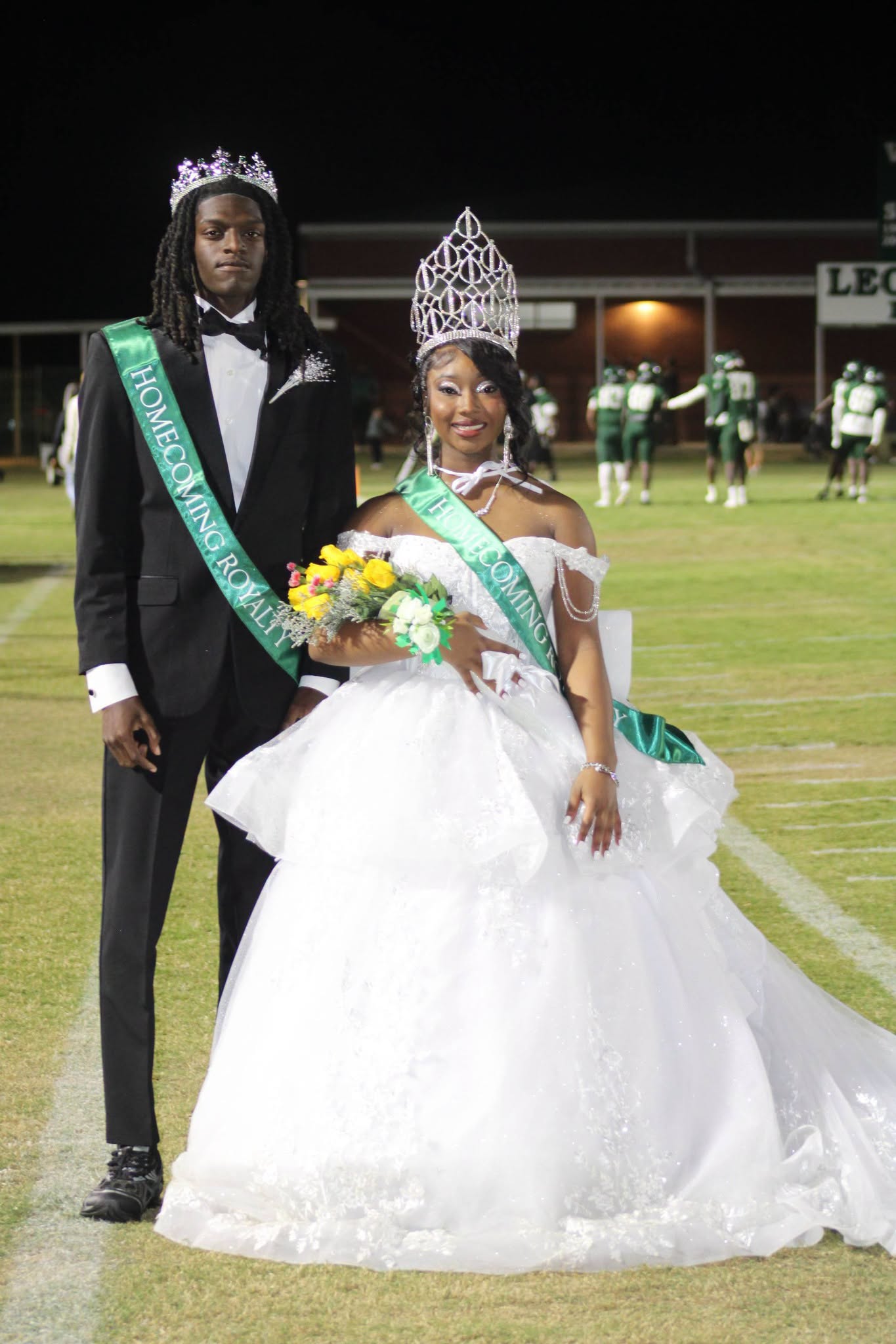 WPHS 2026–2027 Homecoming King Jayden Randle and Homecoming Queen JaKayla Buchanan pose together on the football field wearing crowns and green Homecoming Royalty sashes.